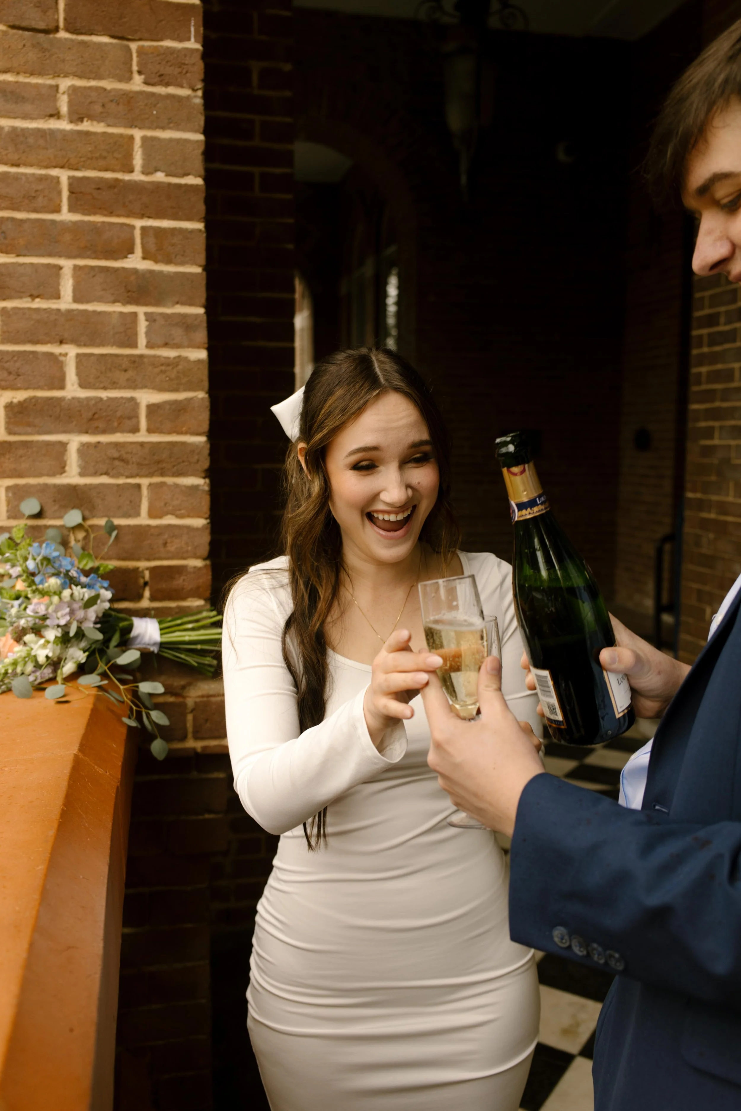 The bride lights up with joy as the groom pours champagne into her glass, bouquet resting on the brick ledge beside them.
