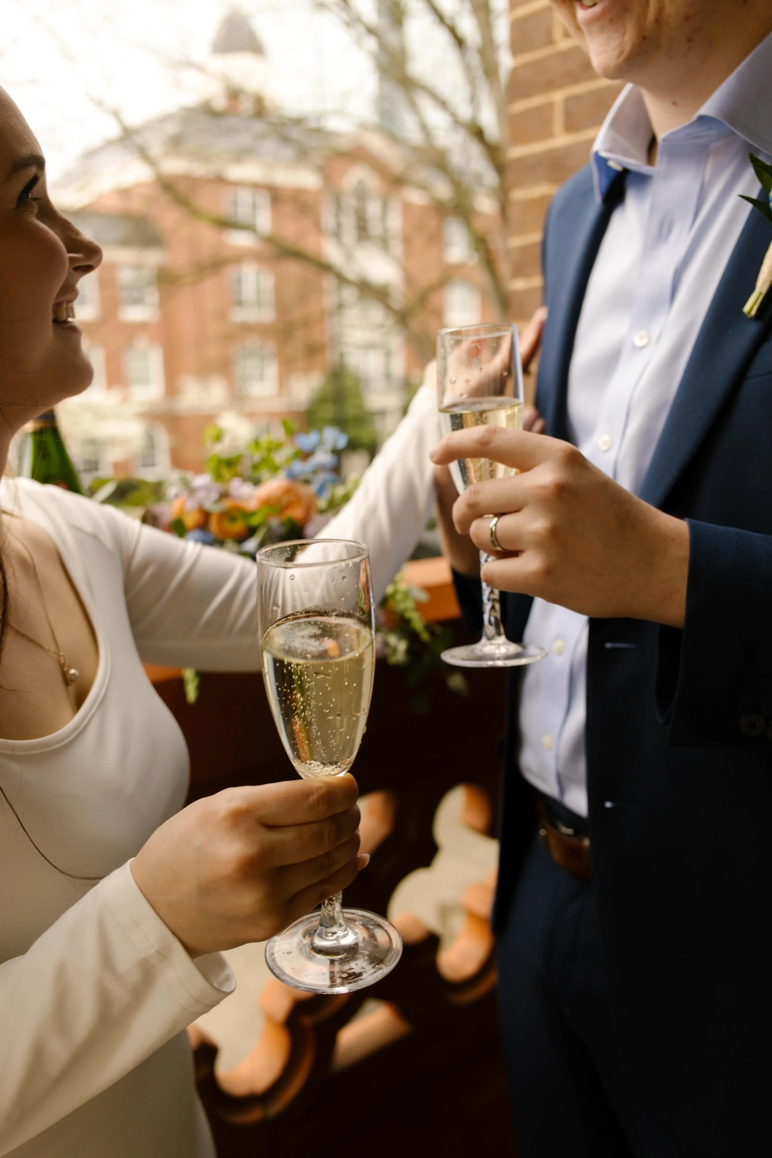 Close-up of the couple clinking champagne glasses, with wedding bands, florals, and the courthouse architecture subtly visible in the background.