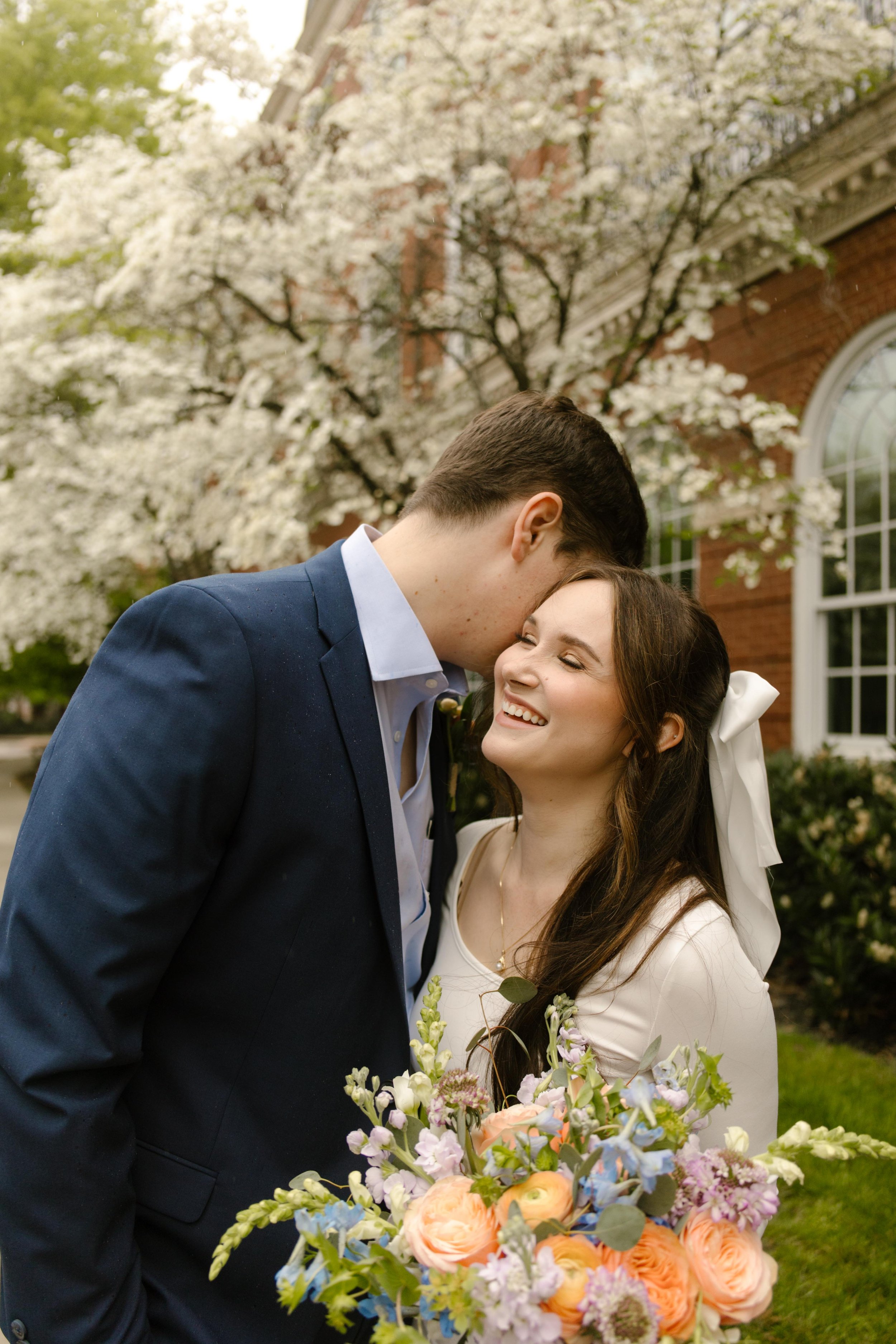 The couple smiles beneath blooming white dogwoods, the bride holding her bouquet as the groom whispers into her ear.