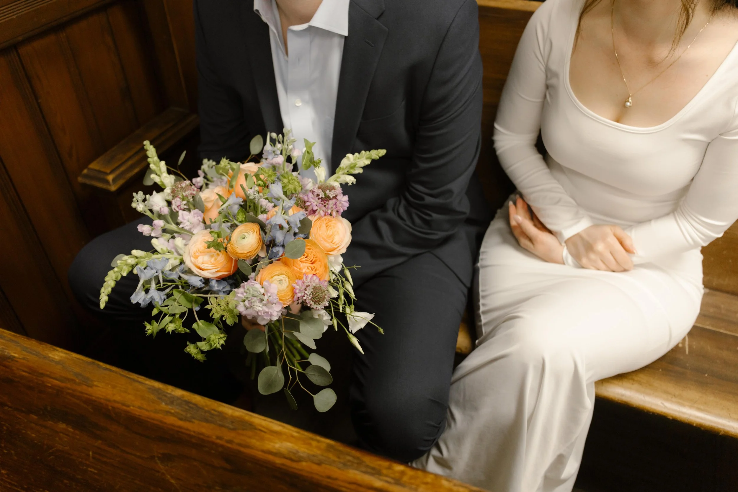 A colorful wedding bouquet rests on the groom’s lap as the couple sits close together on a wooden bench inside the courthouse.