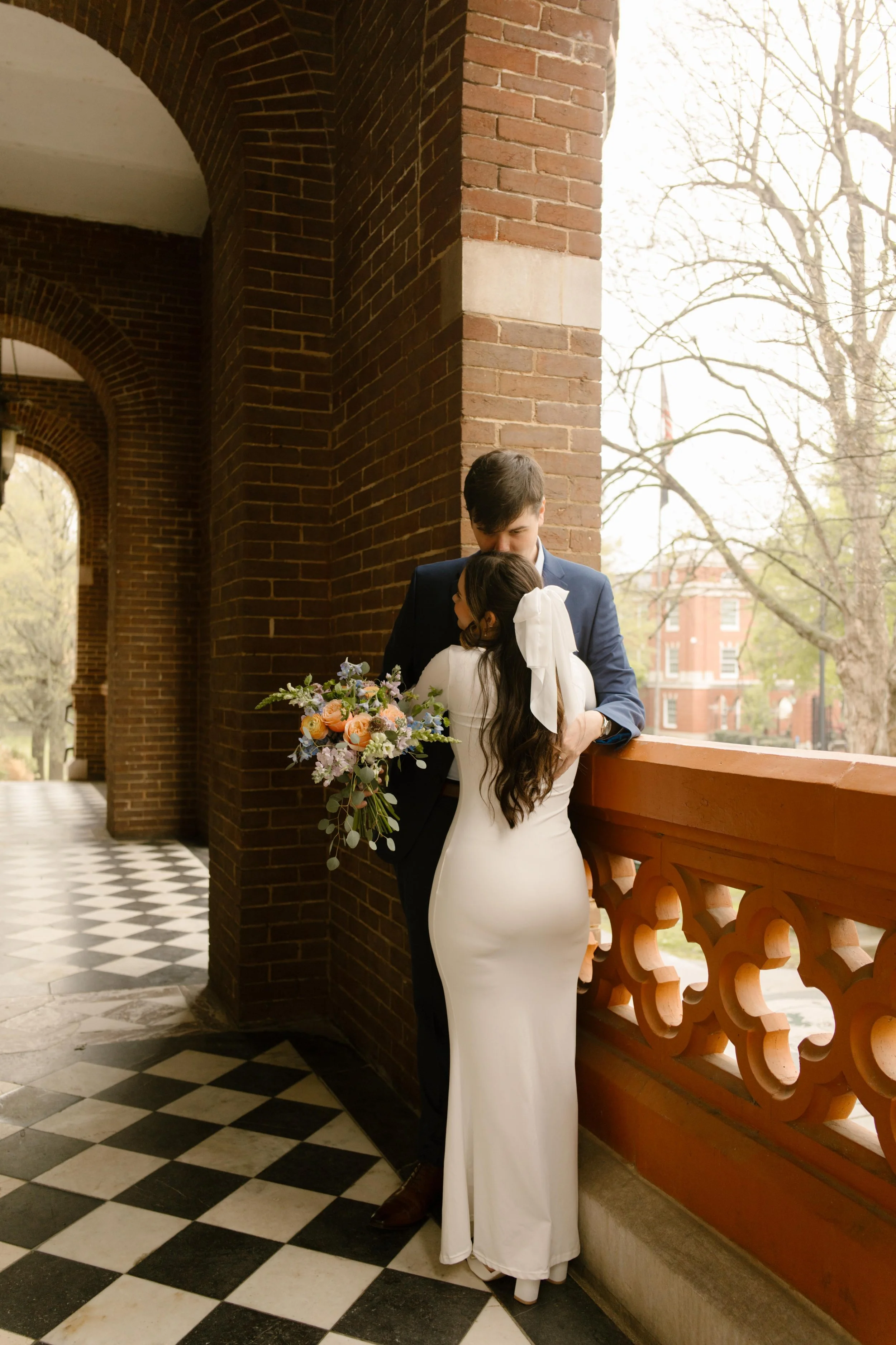 The couple shares a quiet moment on the courthouse balcony, arms wrapped around each other and bouquet resting at her side.