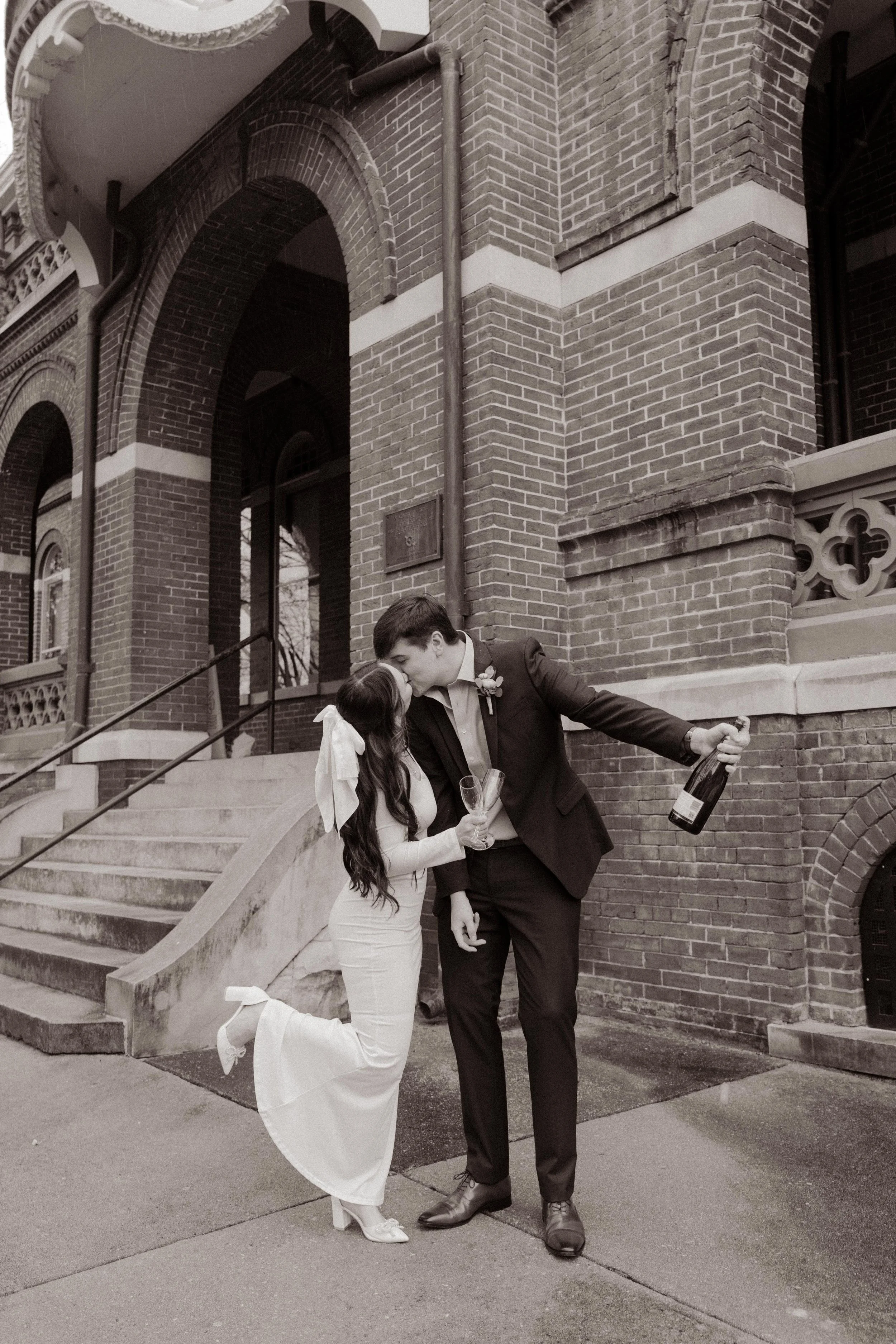 Black-and-white image of the newlyweds sharing a kiss with a champagne bottle in hand outside the historic Knoxville courthouse.