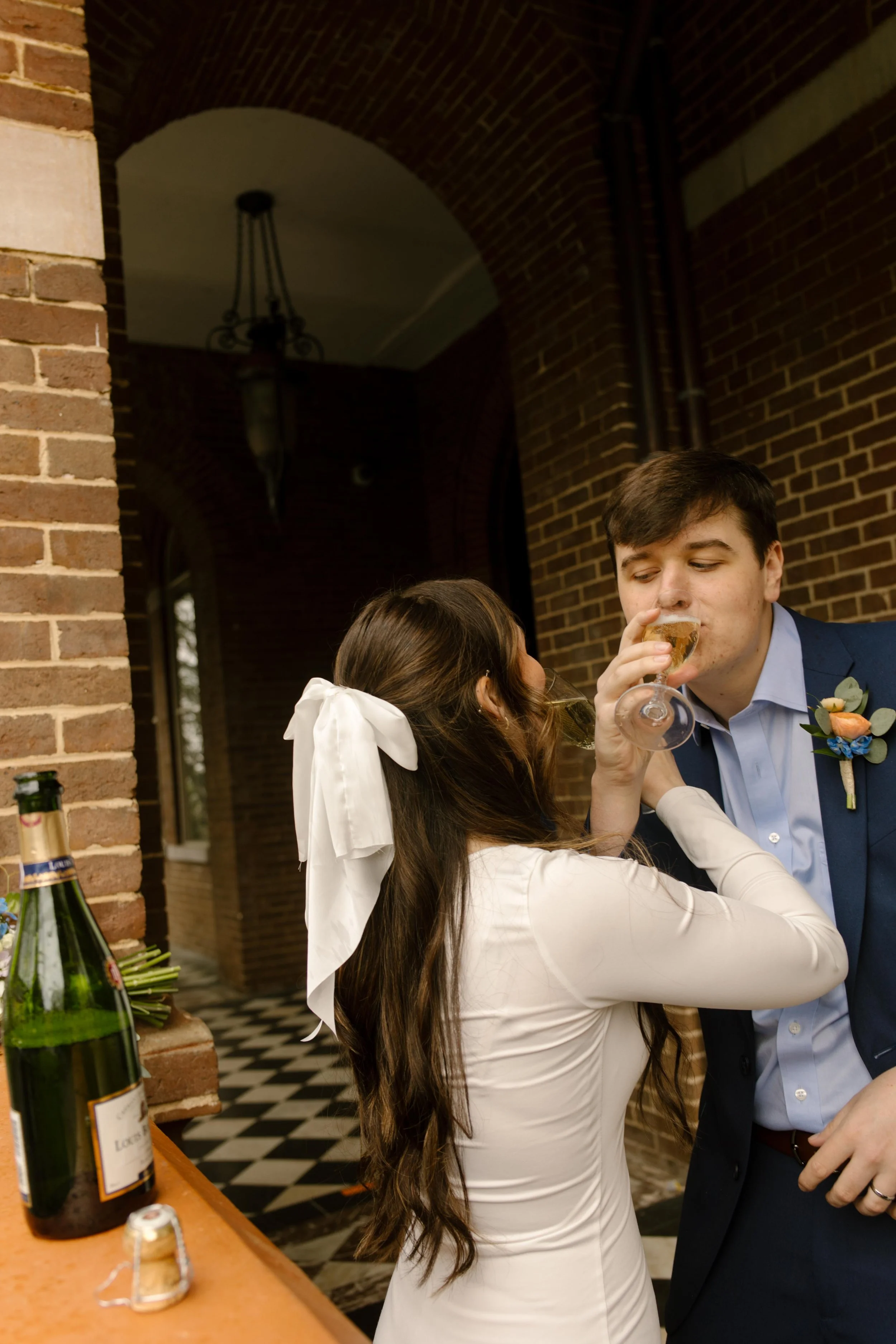 The bride helps her new husband sip champagne as they laugh together against the red brick—celebration in full swing after their Knoxville courthouse elopement.