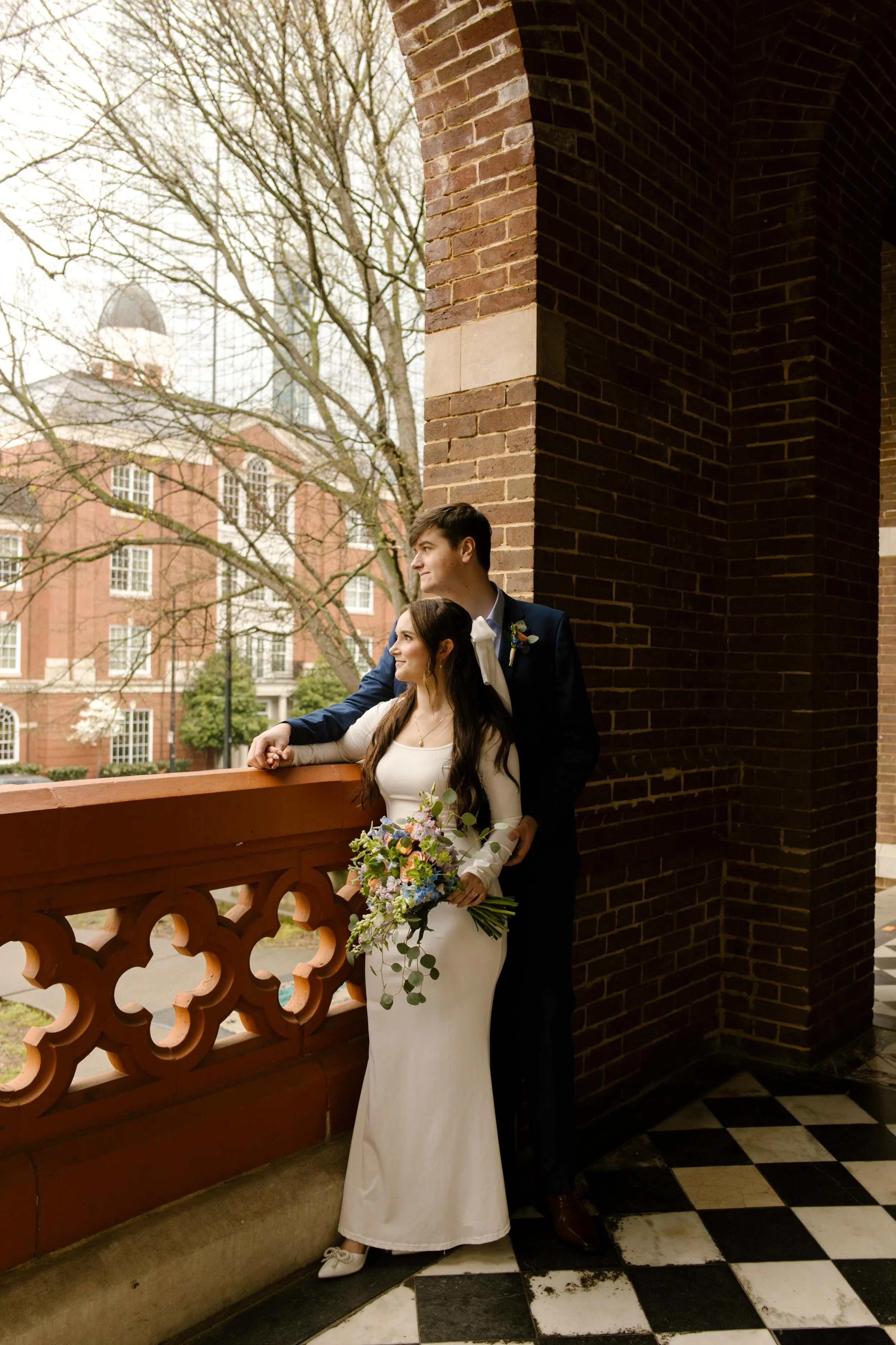 The bride and groom look toward downtown from the courthouse balcony, bouquet in hand, wrapped in the calm of their wedding day.