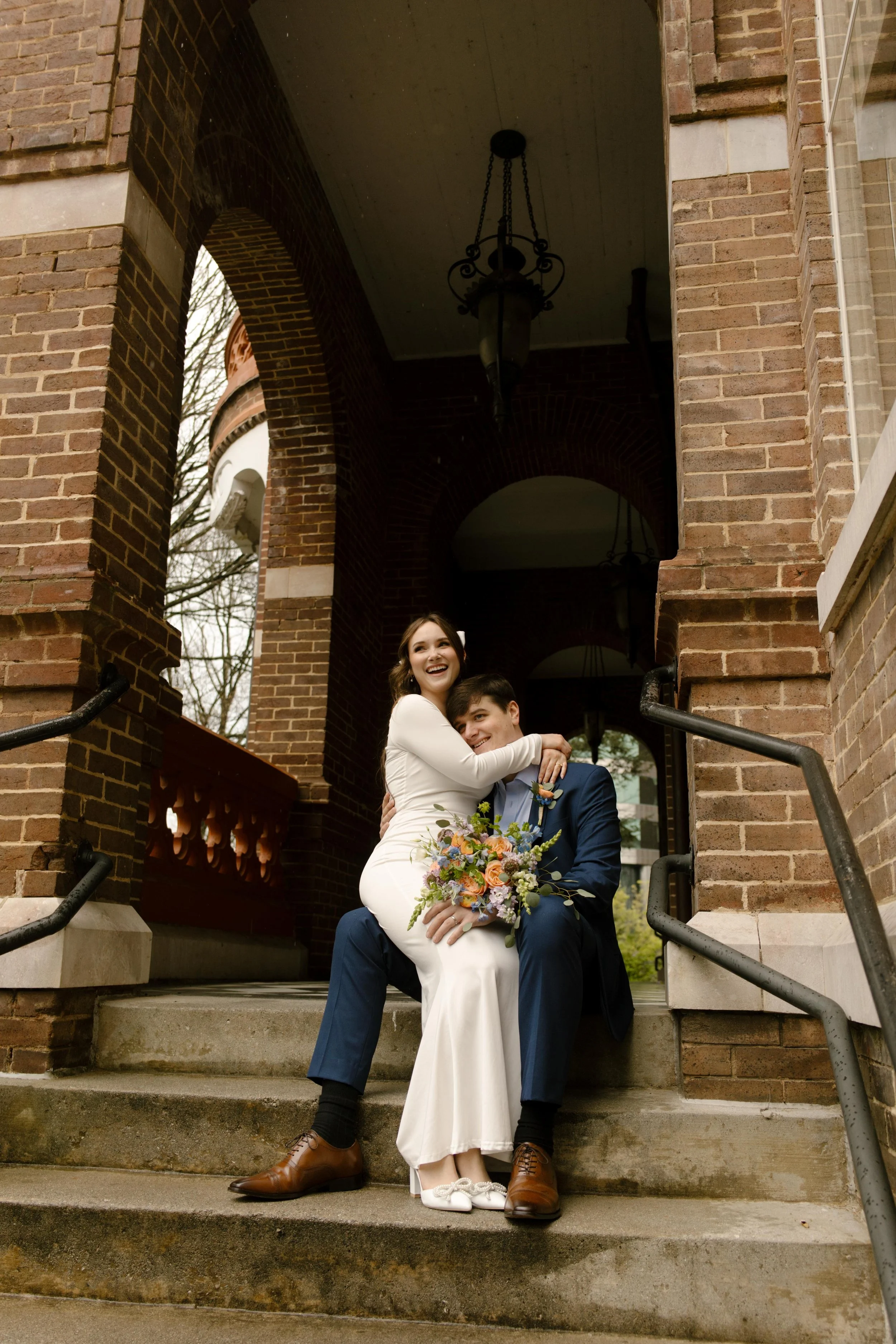 The bride sits on the groom’s lap on the courthouse steps, bouquet resting in her hands and both of them glowing with just-married giddiness.