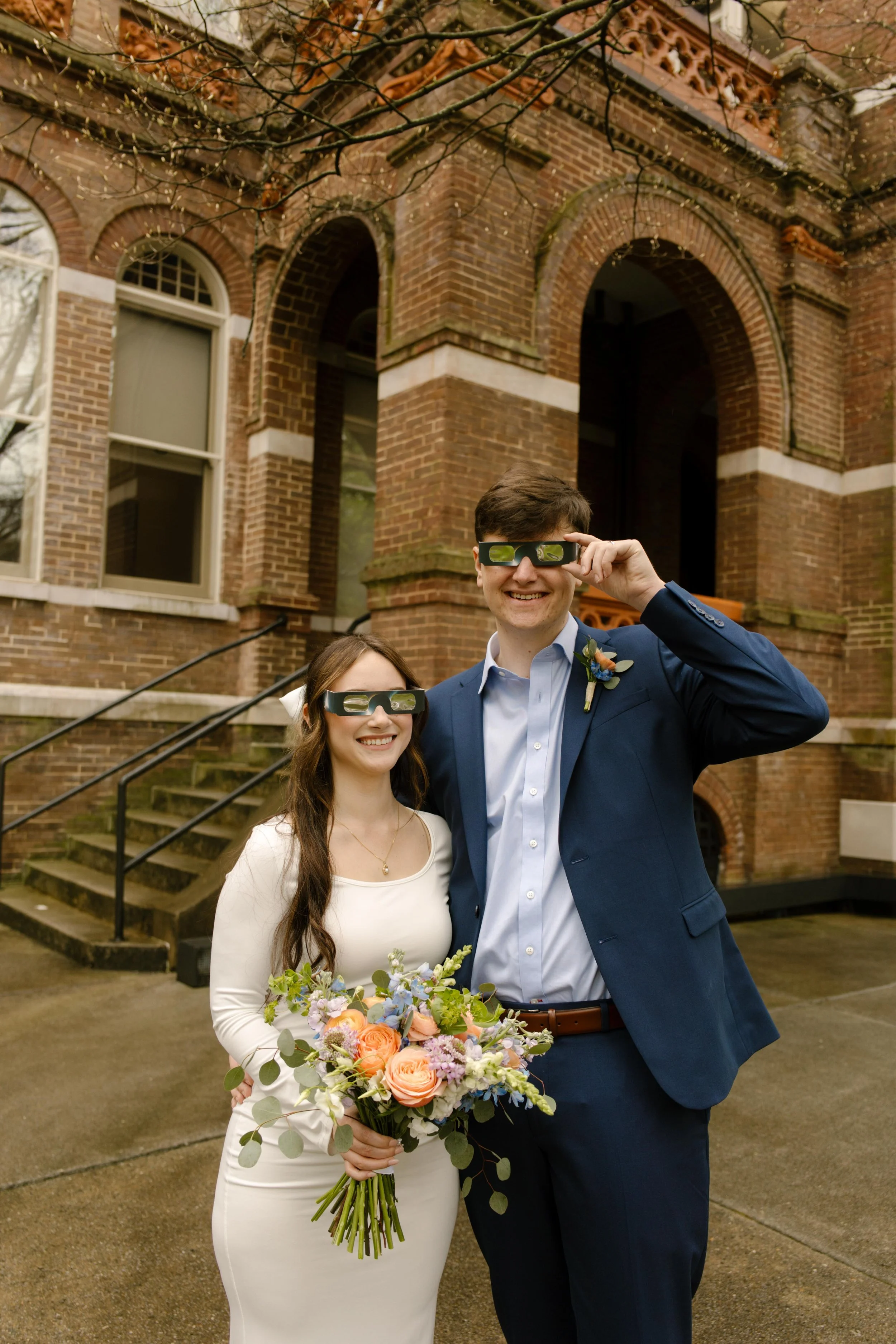 Newlyweds pose with eclipse glasses and wildflower bouquet outside the courthouse—sunny smiles and celestial vibes all around.