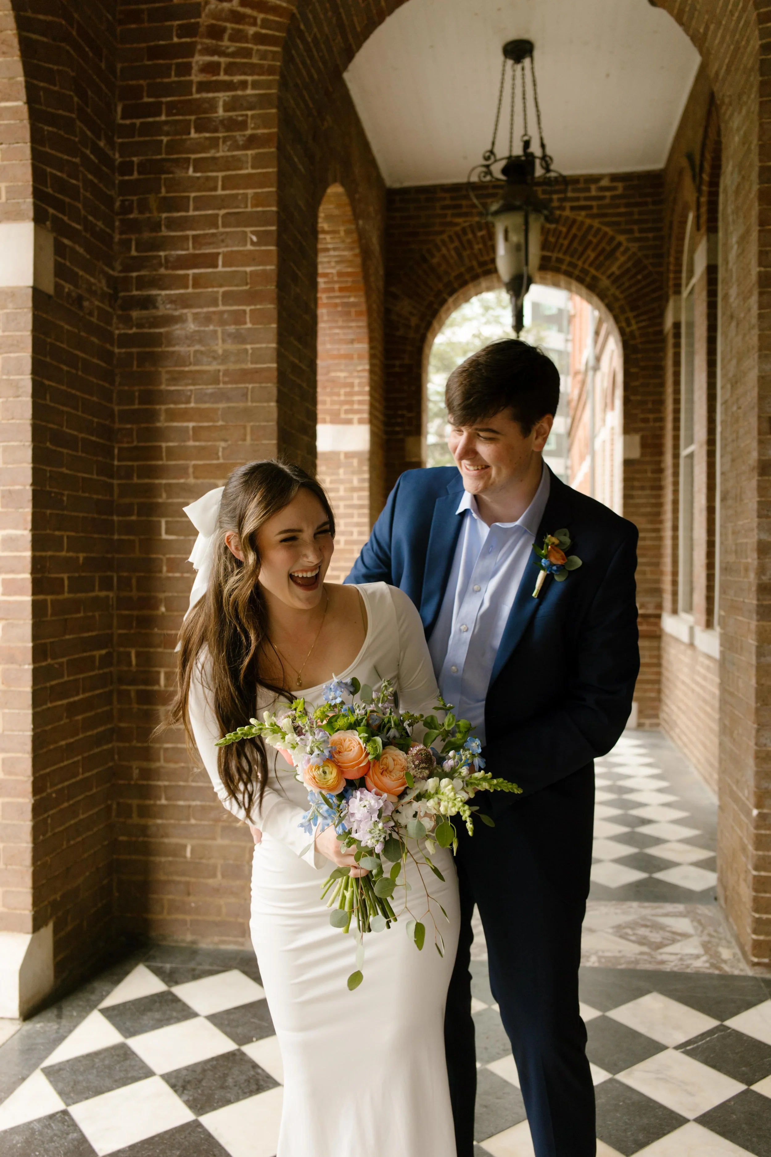 In a moment of playful flirtation, the groom wraps his arms around the bride as they laugh beneath the courthouse arches.