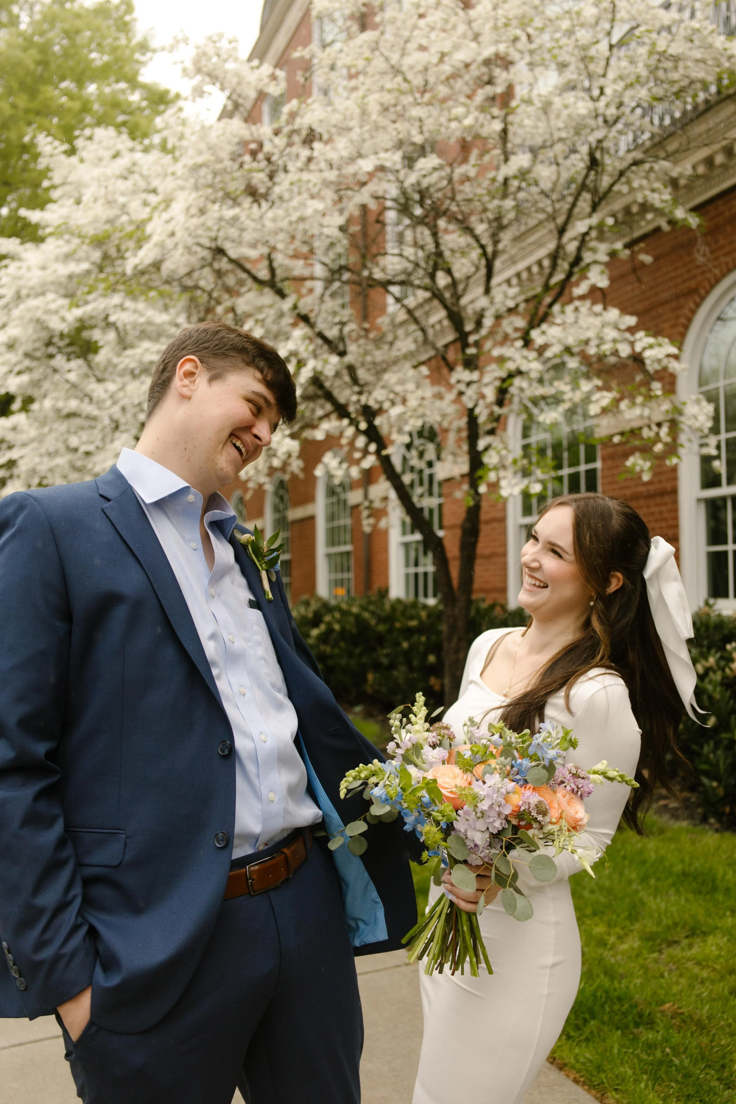 The couple shares a joyful laugh under blooming dogwoods, the bride holding her pastel bouquet with spring magic in the air.