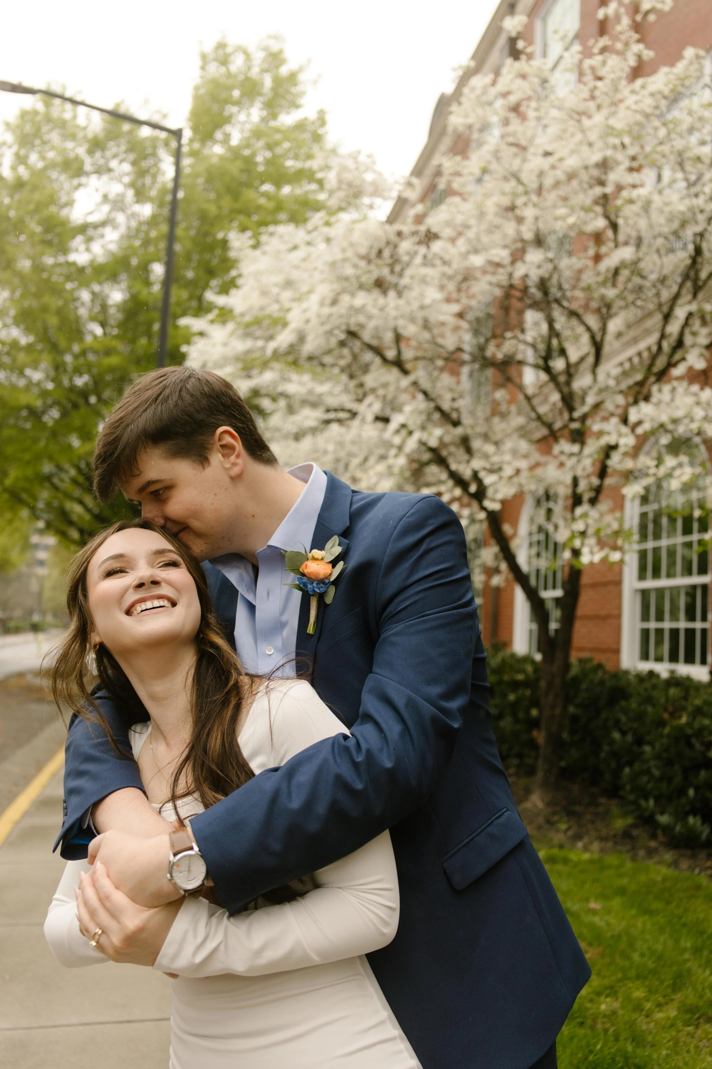 Wrapped in a tight hug on the sidewalk, the groom kisses the bride’s cheek as she smiles, framed by flowering trees and post-ceremony bliss.