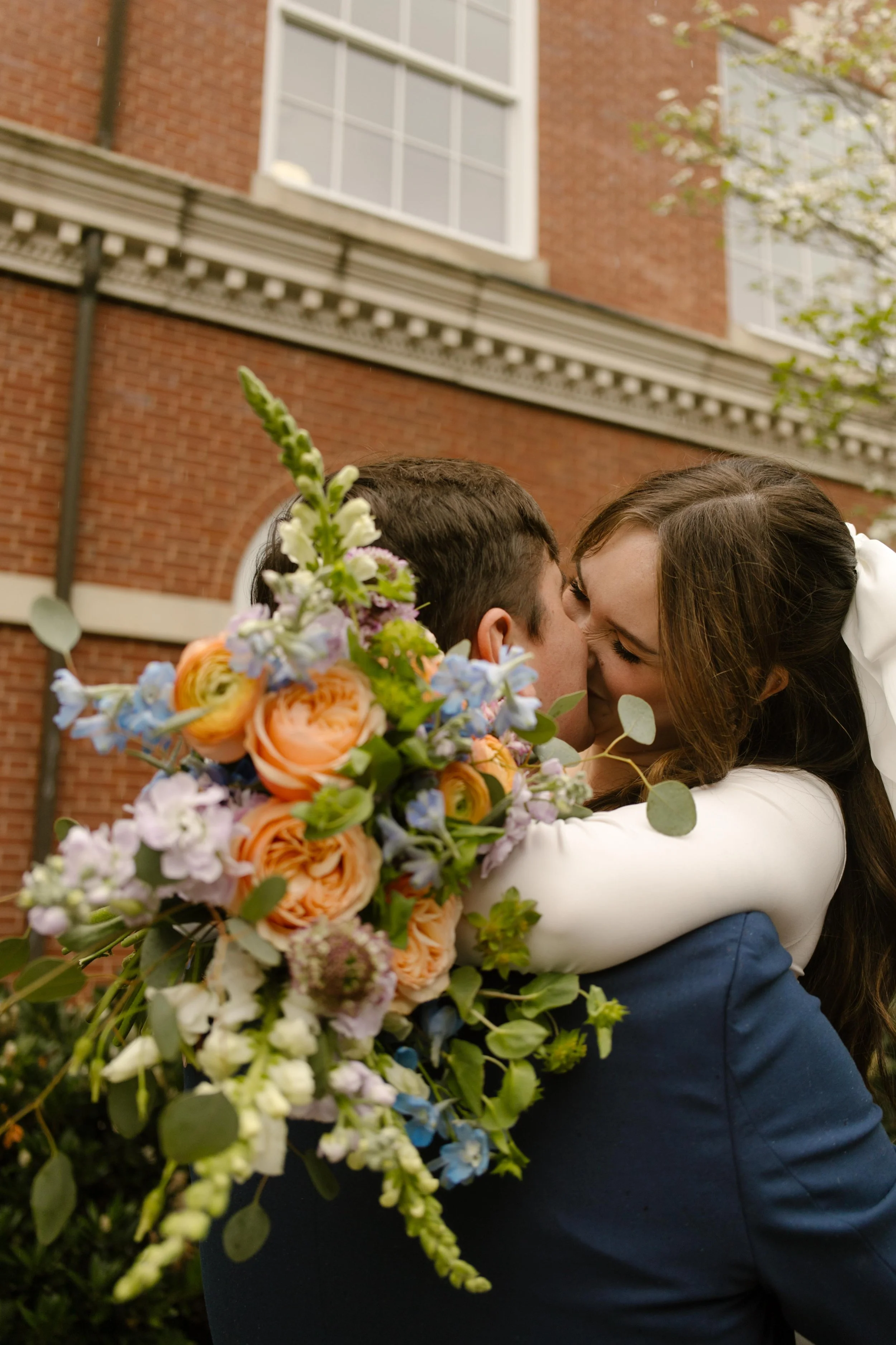 Close-up of the bride smiling into a kiss, her bouquet of ranunculus and wildflowers taking center stage against the groom’s shoulder.