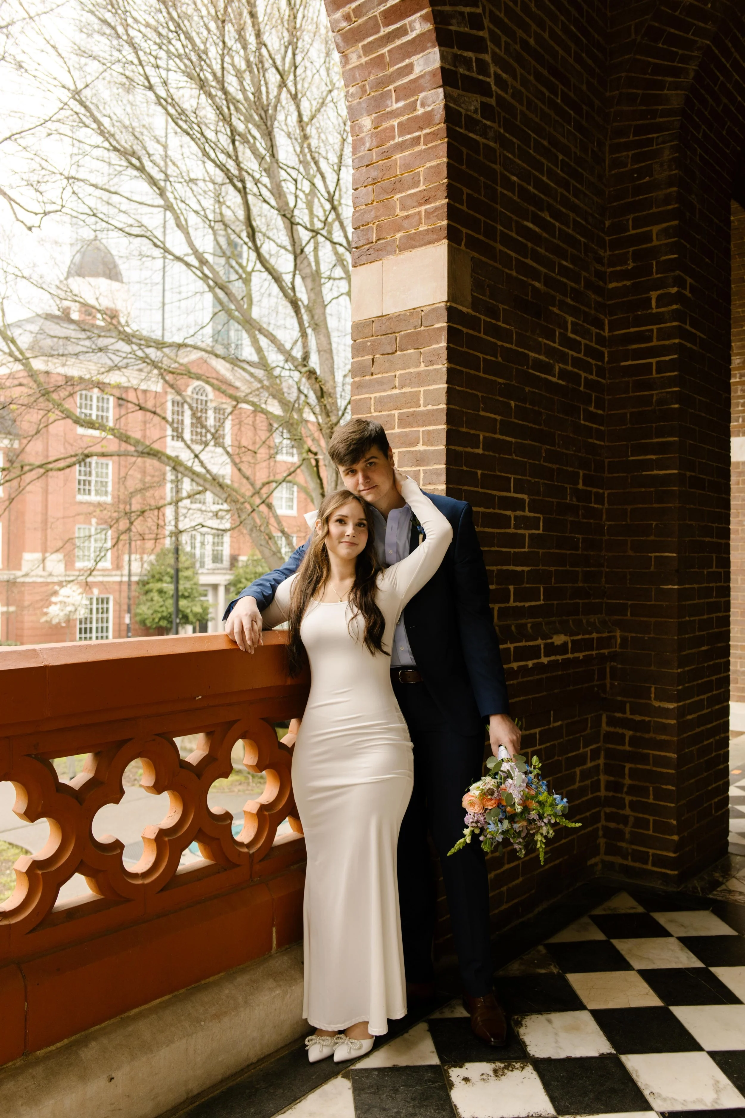 With a bouquet in one hand and her arm draped around his shoulder, the bride leans into her groom as they pose on the courthouse balcony after their Knoxville courthouse elopement.