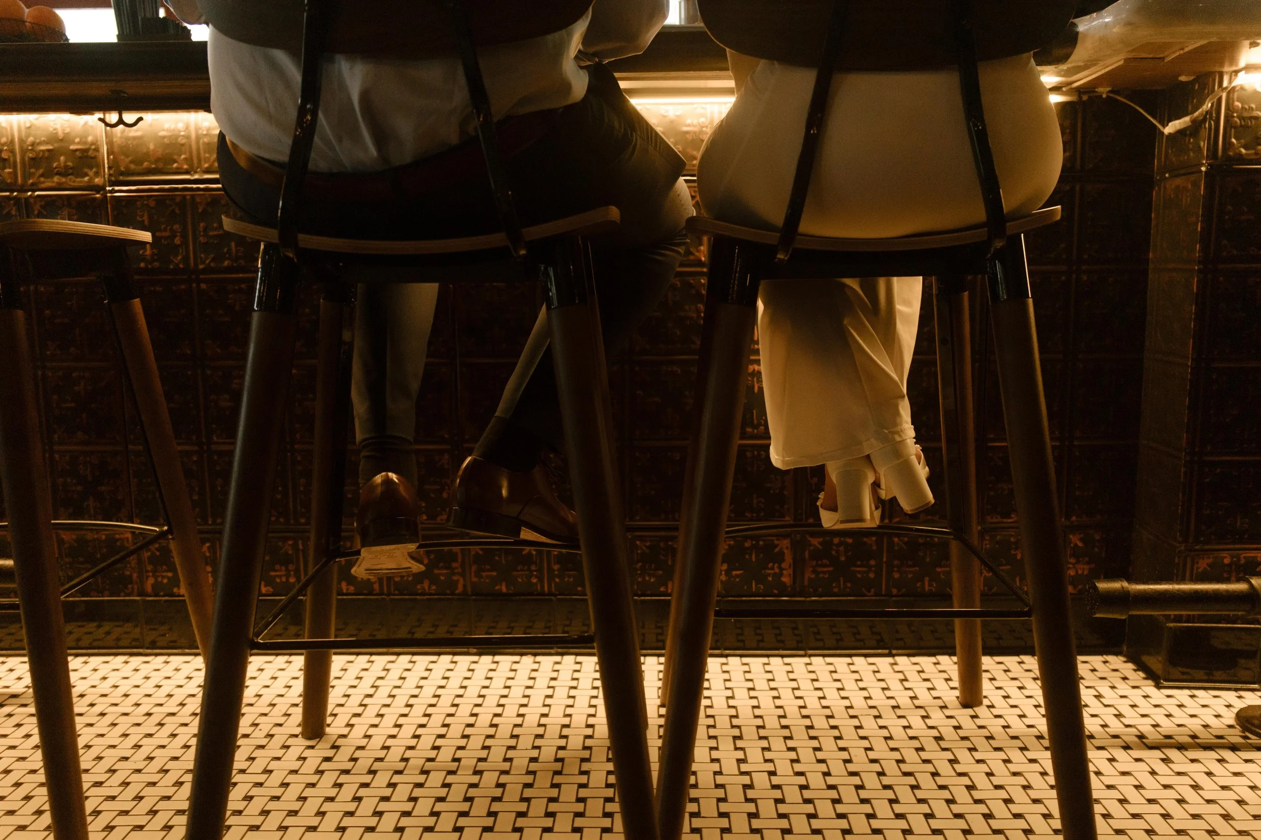 Shot from behind, the couple sits at the bar, their feet just barely touching the floor—still in wedding clothes but deep in their own little world.