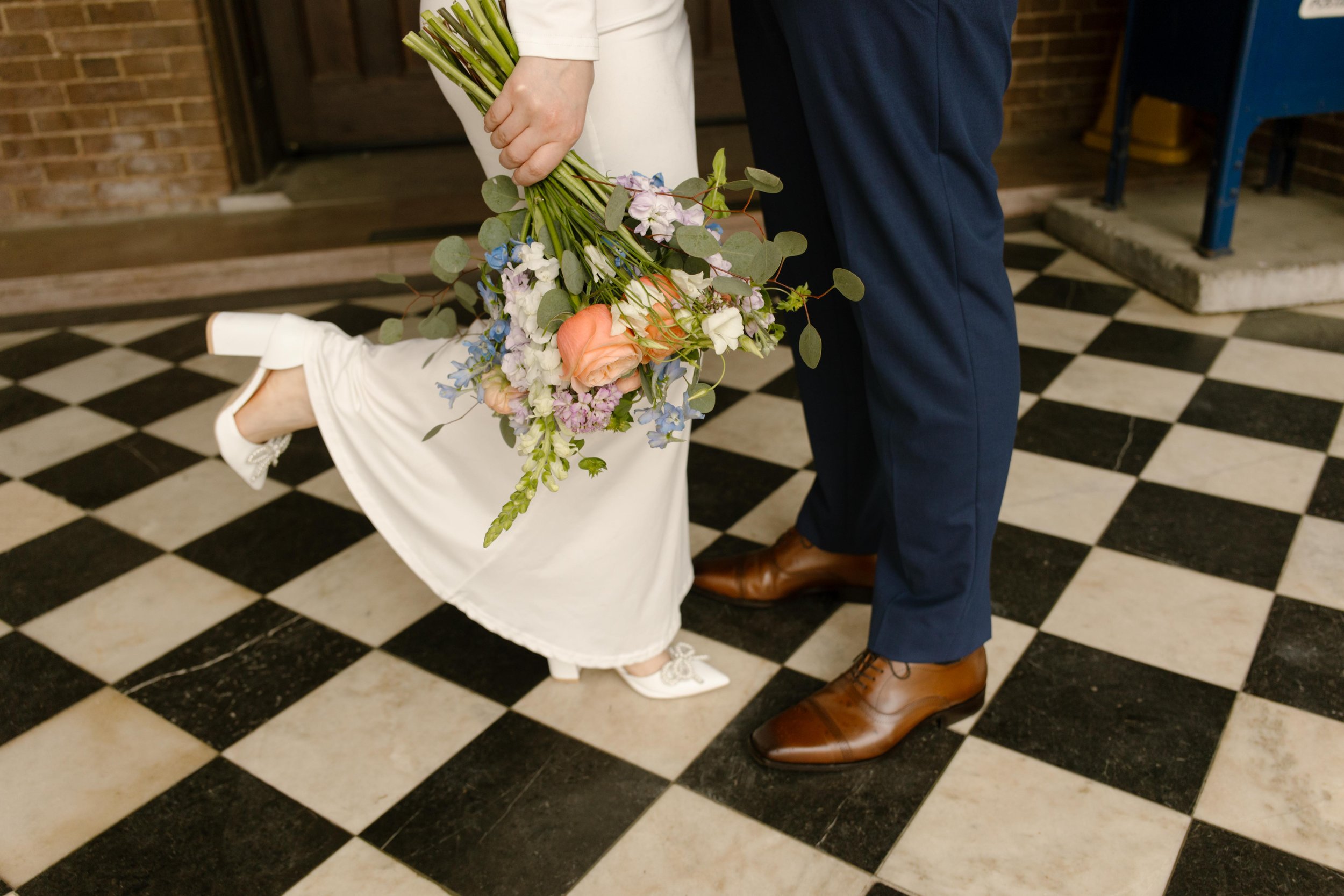 A playful detail shot of their shoes and intertwined legs on the courthouse tiles—bouquet dangling, one heel kicked up mid-romance.
