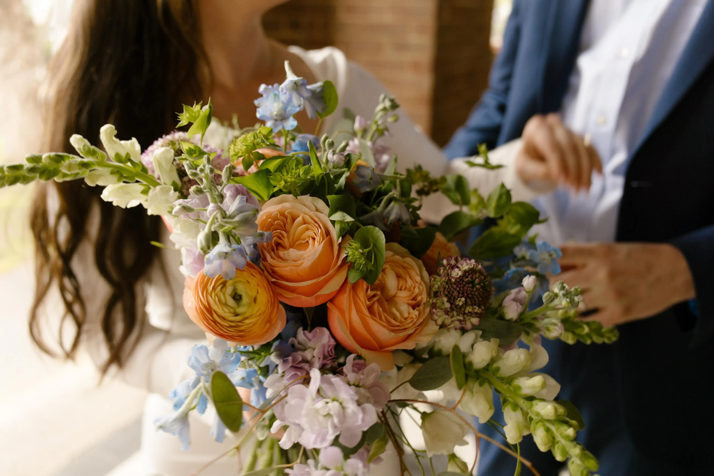 A vibrant close-up of the bride’s bouquet with apricot ranunculus, blue delphinium, and lilac stems—freshly held outside the courthouse.