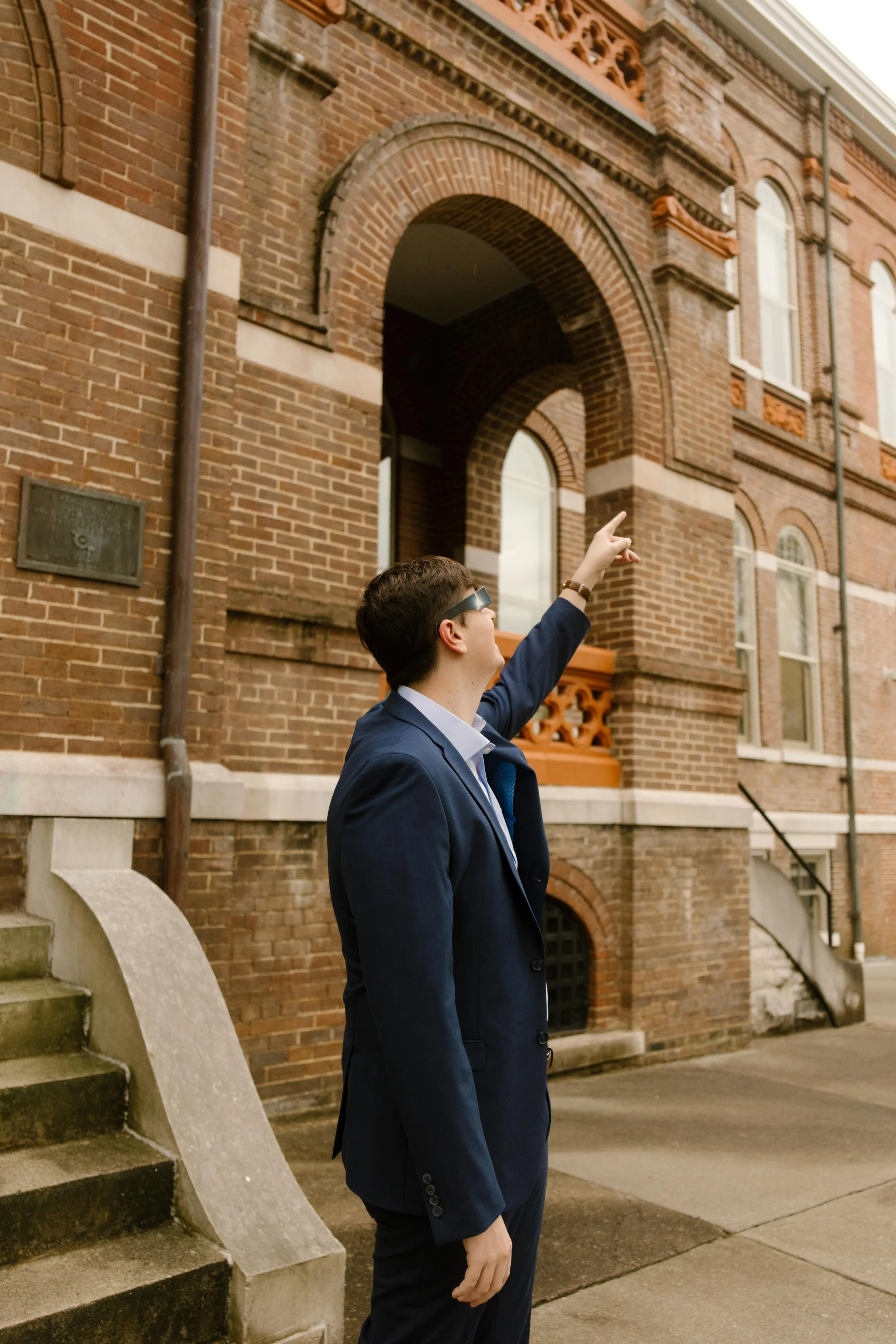 The groom, wearing sunglasses and a sharp navy suit, points up toward the historic brick courthouse, mid-laugh and full of celebration vibes.