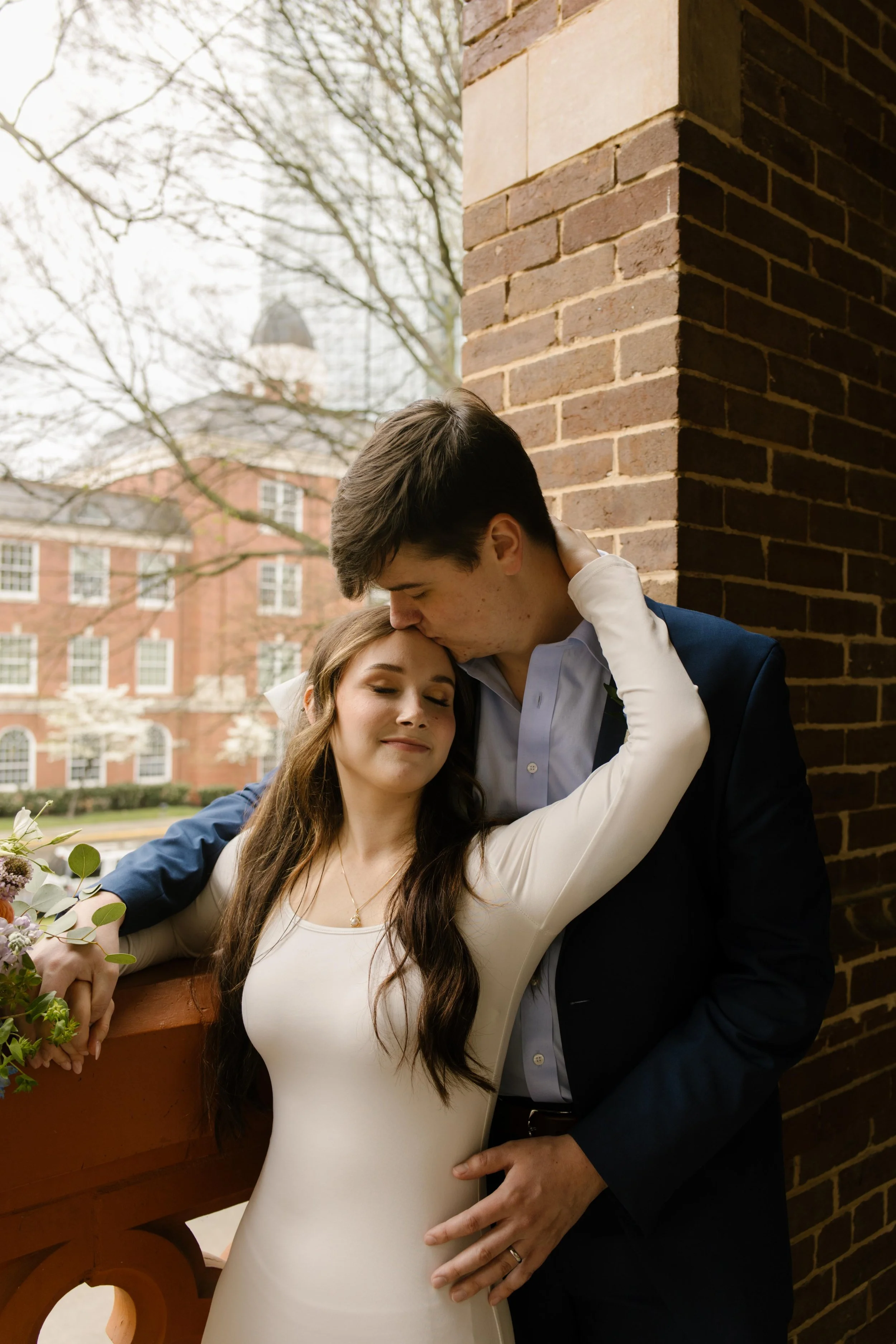 Groom kisses the bride’s forehead as she melts into his embrace, eyes closed, on the courthouse balcony—pure tenderness after their Knoxville courthouse elopement.
