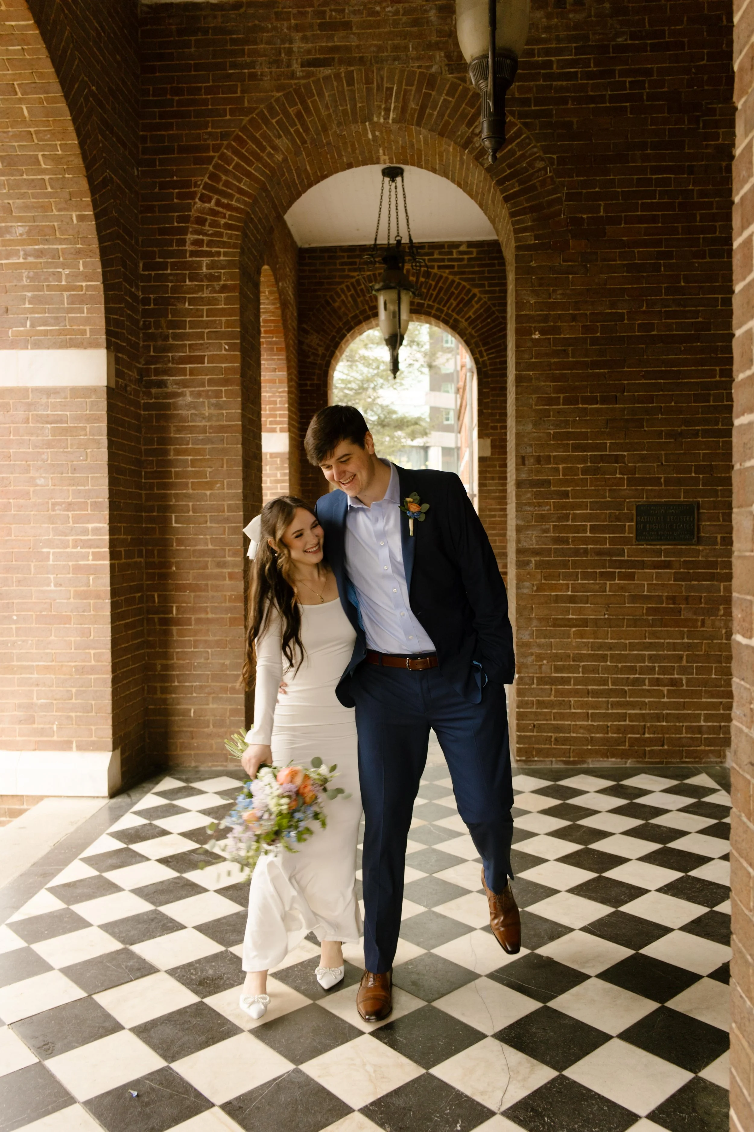 The couple shares a quiet laugh, walking arm-in-arm beneath the courthouse arches, bouquet swinging between them like a secret.
