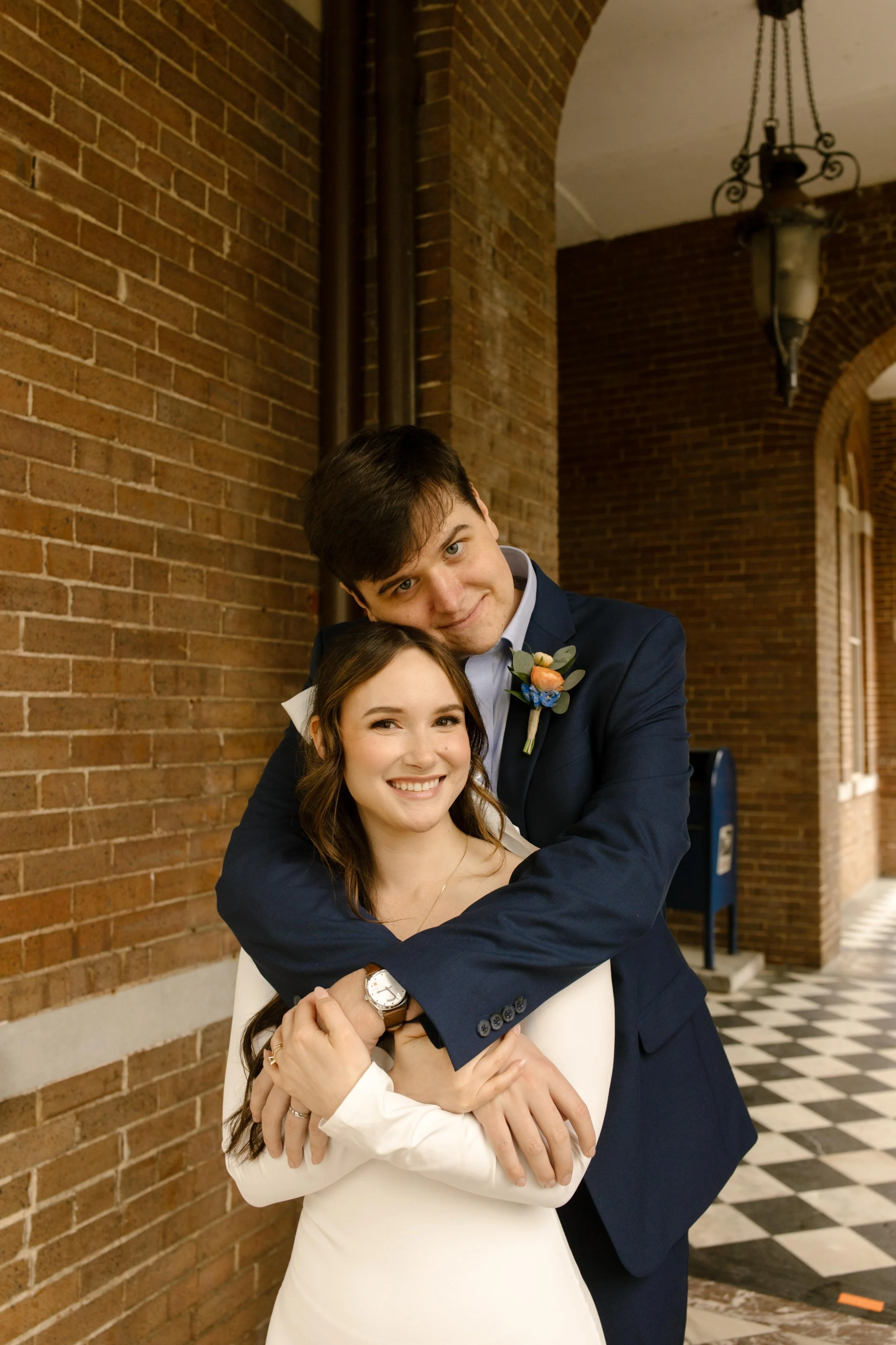 Groom wraps the bride in a tight hug from behind as they smile under the courthouse arches, totally in sync and glowing.