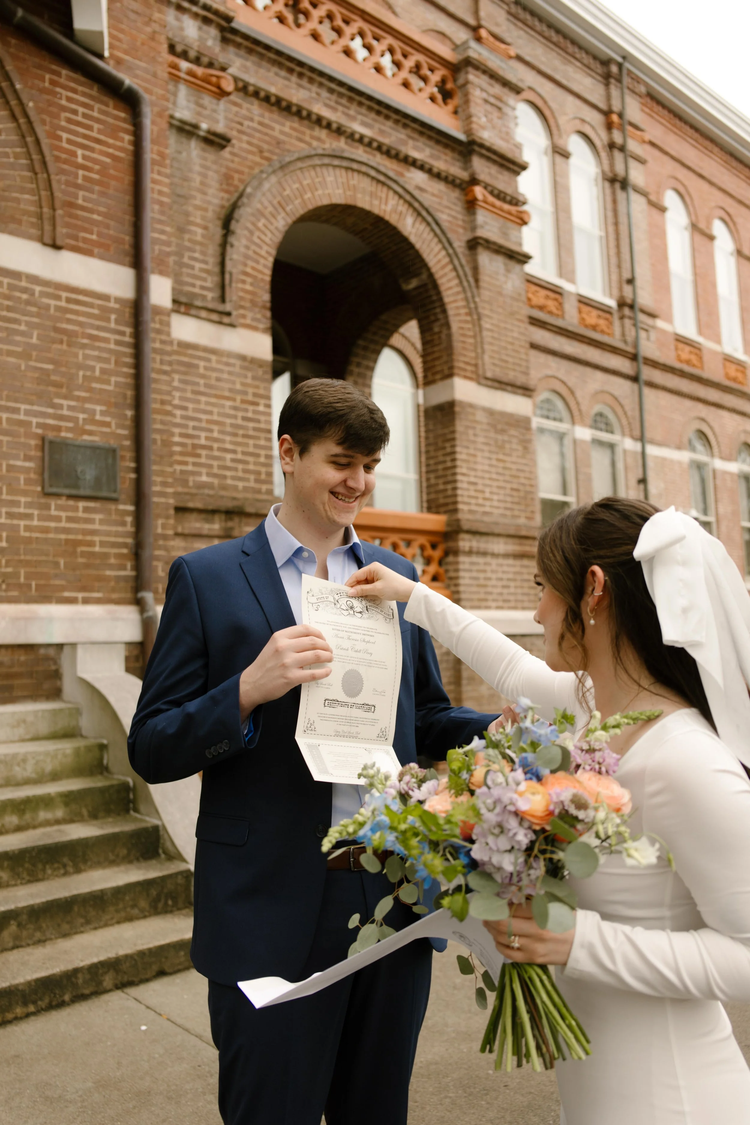 The couple shares a playful moment outside the courthouse as the groom proudly holds their marriage certificate—bouquet in hand, joy in the air after their Knoxville courthouse elopement.