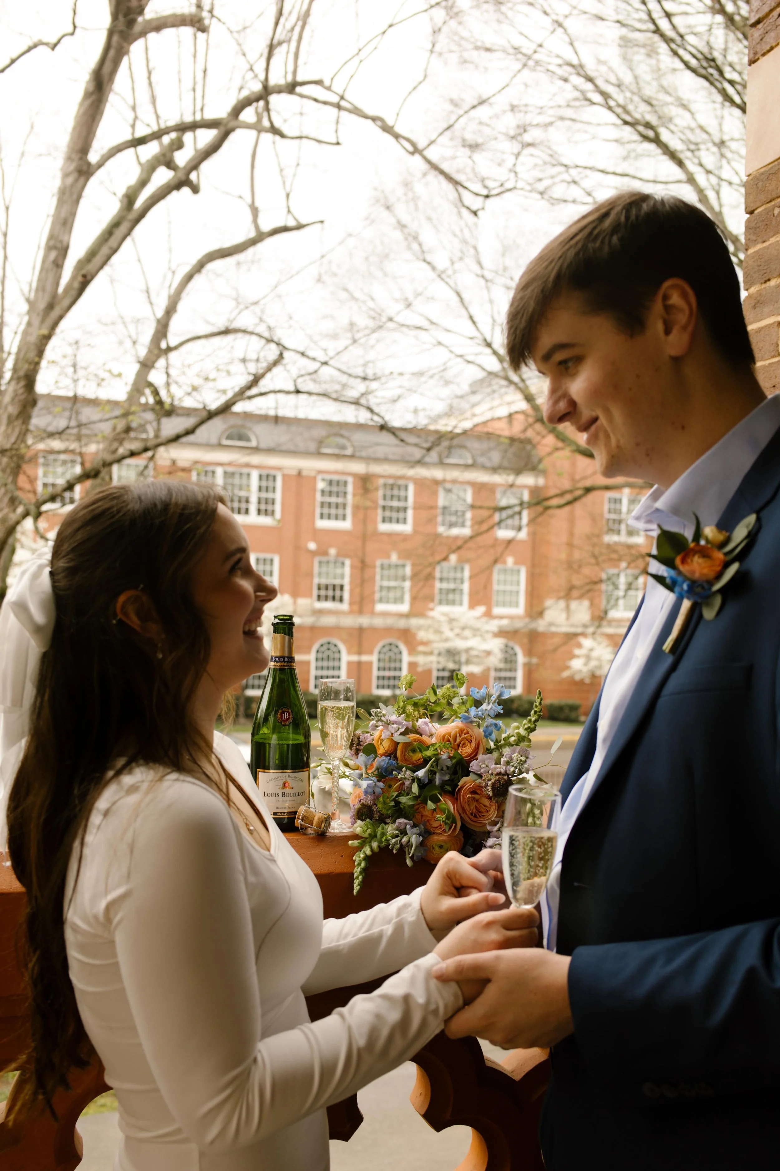 Champagne glasses in hand, the couple locks eyes and shares a smile beside their floral-draped setup on the courthouse balcony—just married and soaking it in.