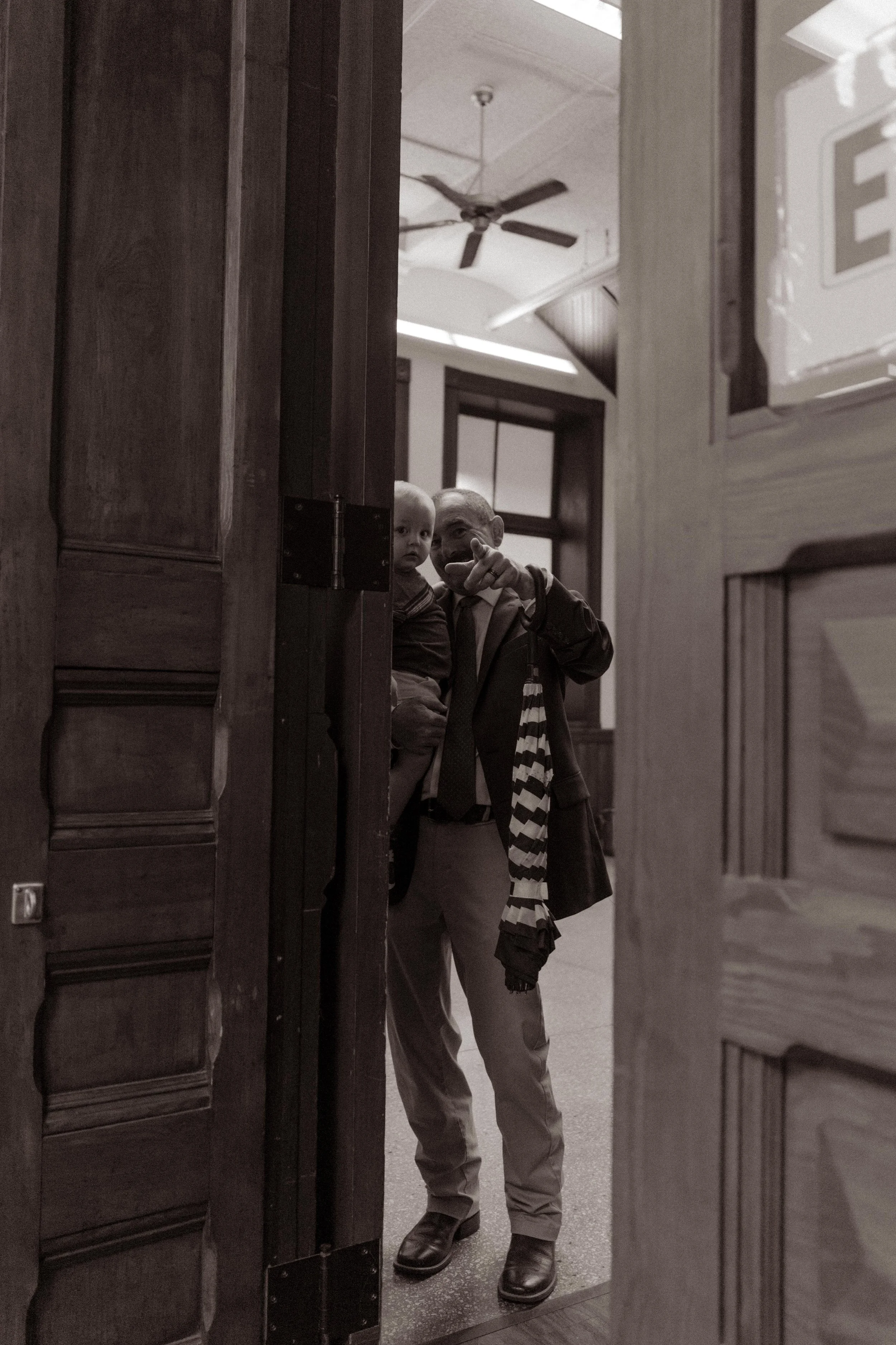 A candid black-and-white photo of a proud grandfather holding a toddler as they peek through the courthouse door with big smiles and waves.