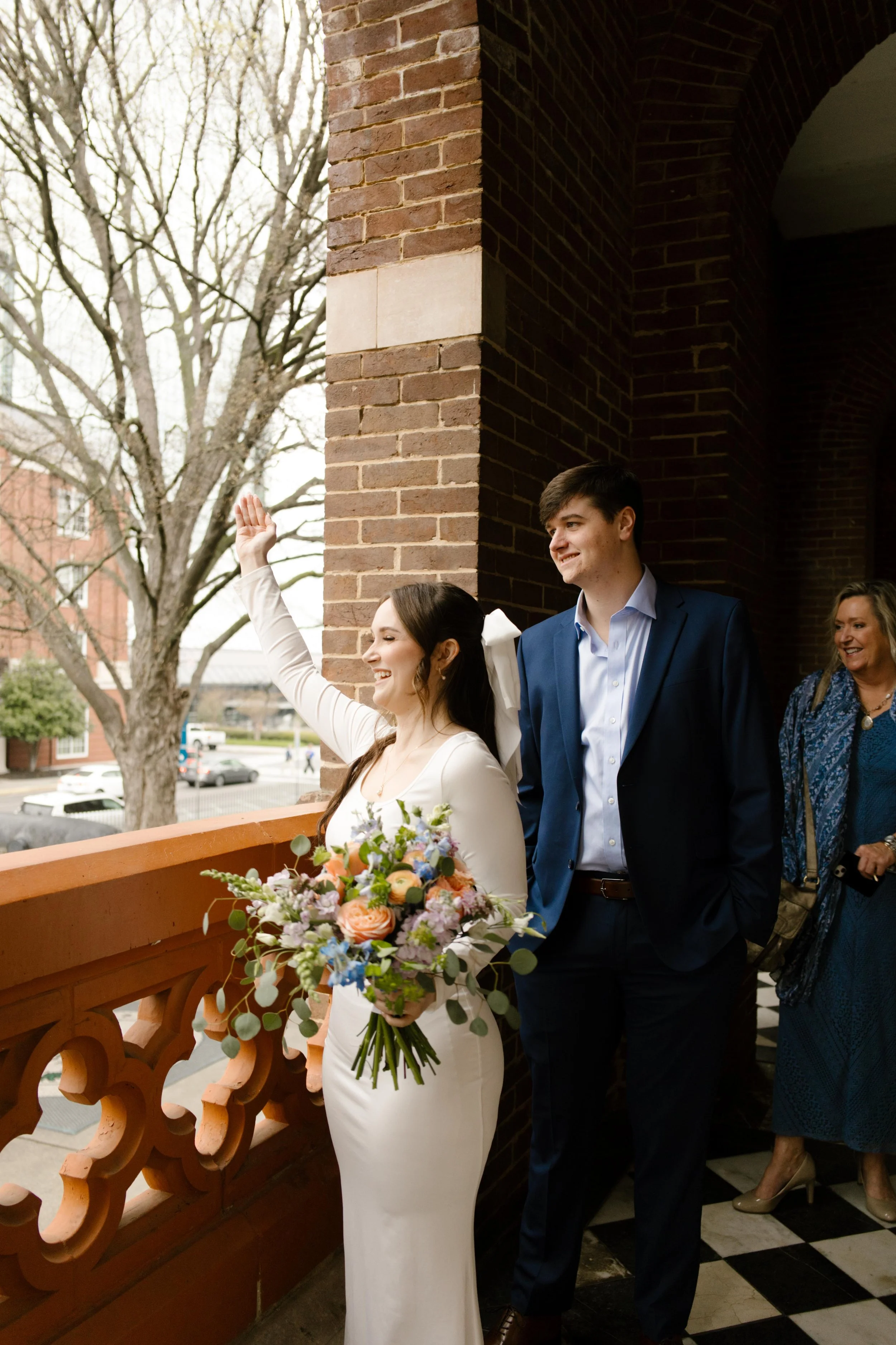 Bride waves joyfully from the brick balcony of the courthouse, bouquet in hand, surrounded by her groom and loved ones during their Knoxville courthouse elopement.