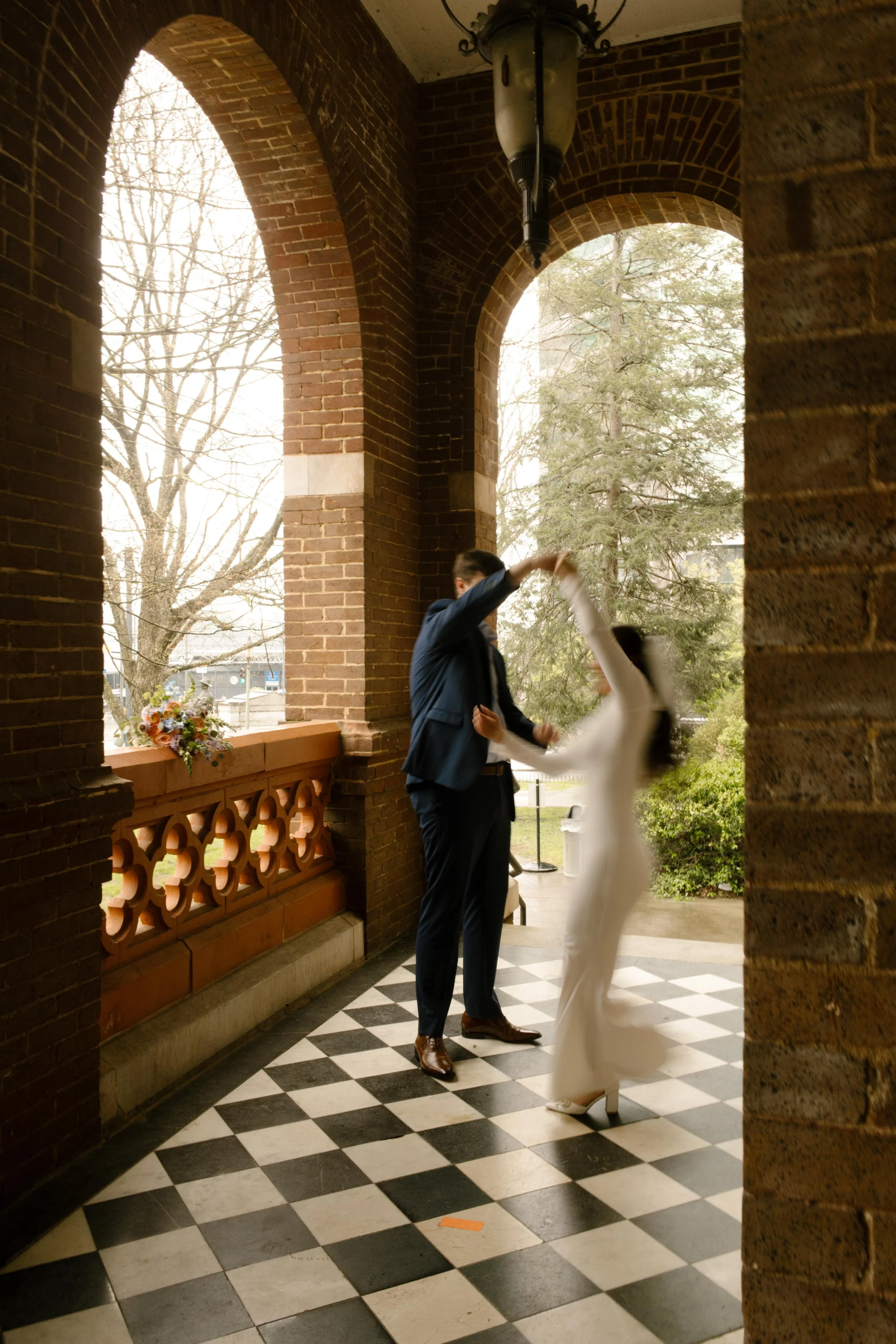 The groom twirls the bride on the checkered courthouse floor, mid-spin and full of joy beneath the brick arches.
