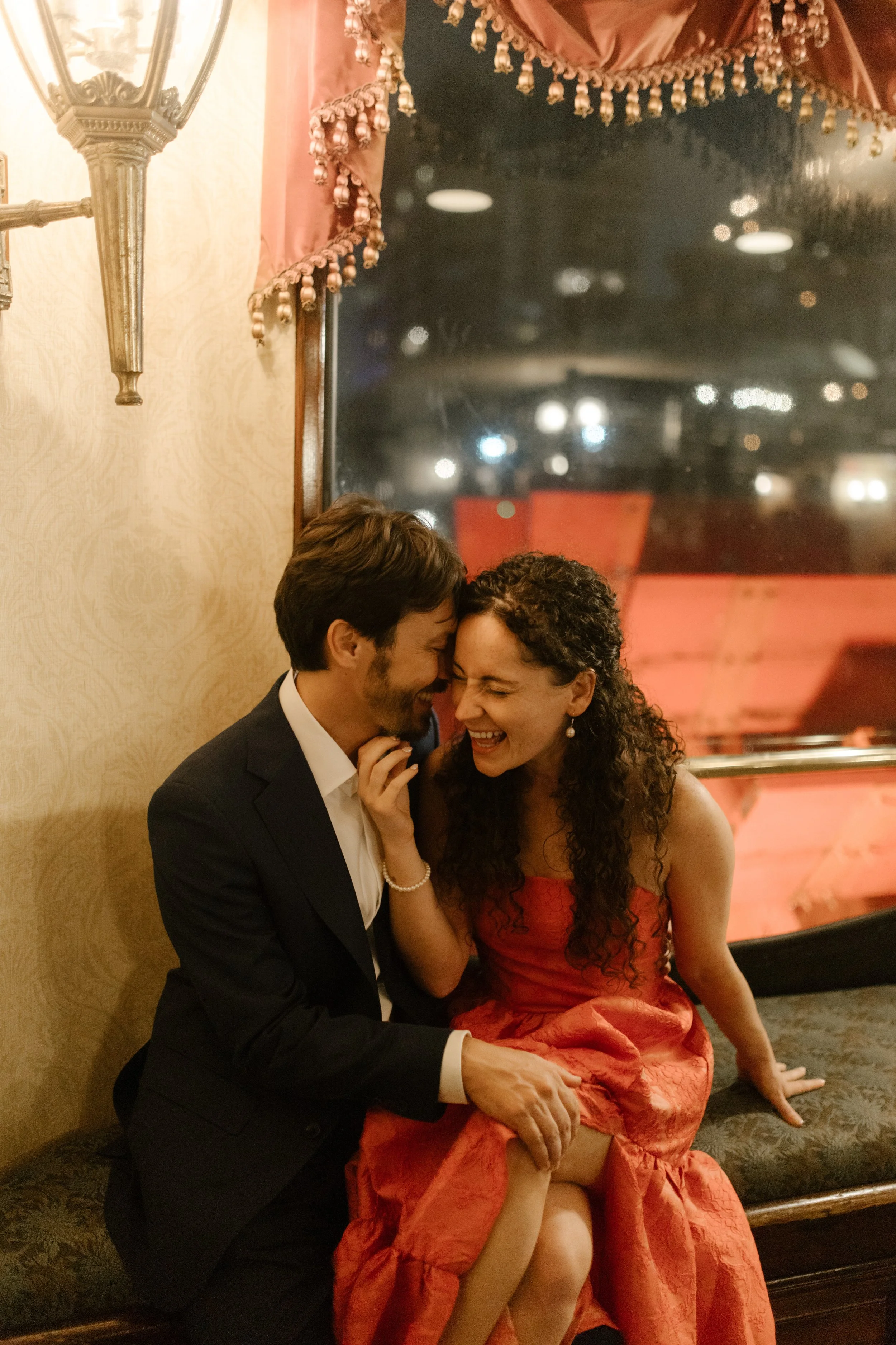 Couple sharing a sweet, private moment indoors—whispering and laughing in the golden glow of their wedding night.