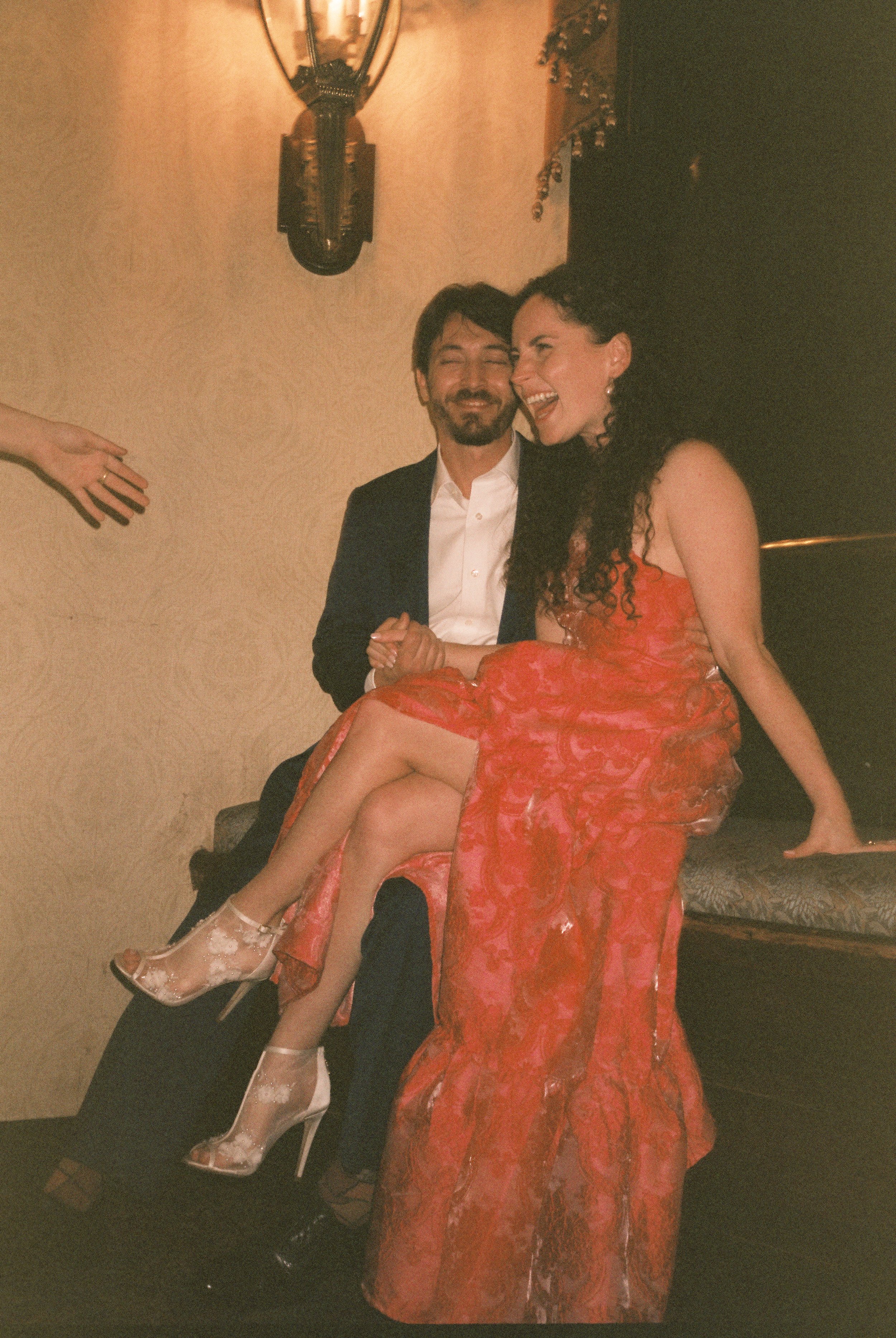Candid flash photo of the couple laughing and cozying up on a bench, post-ceremony bliss during their wedding weekend.