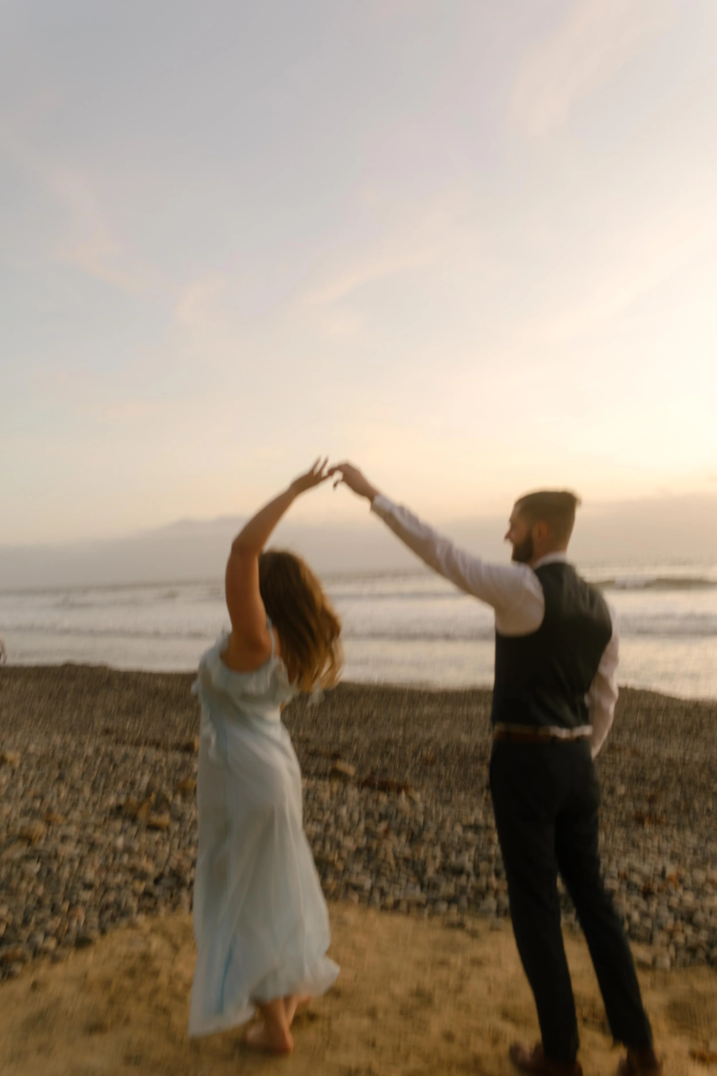 A couple twirls on the shore during sunset, her dress catching the light as waves roll in behind them.