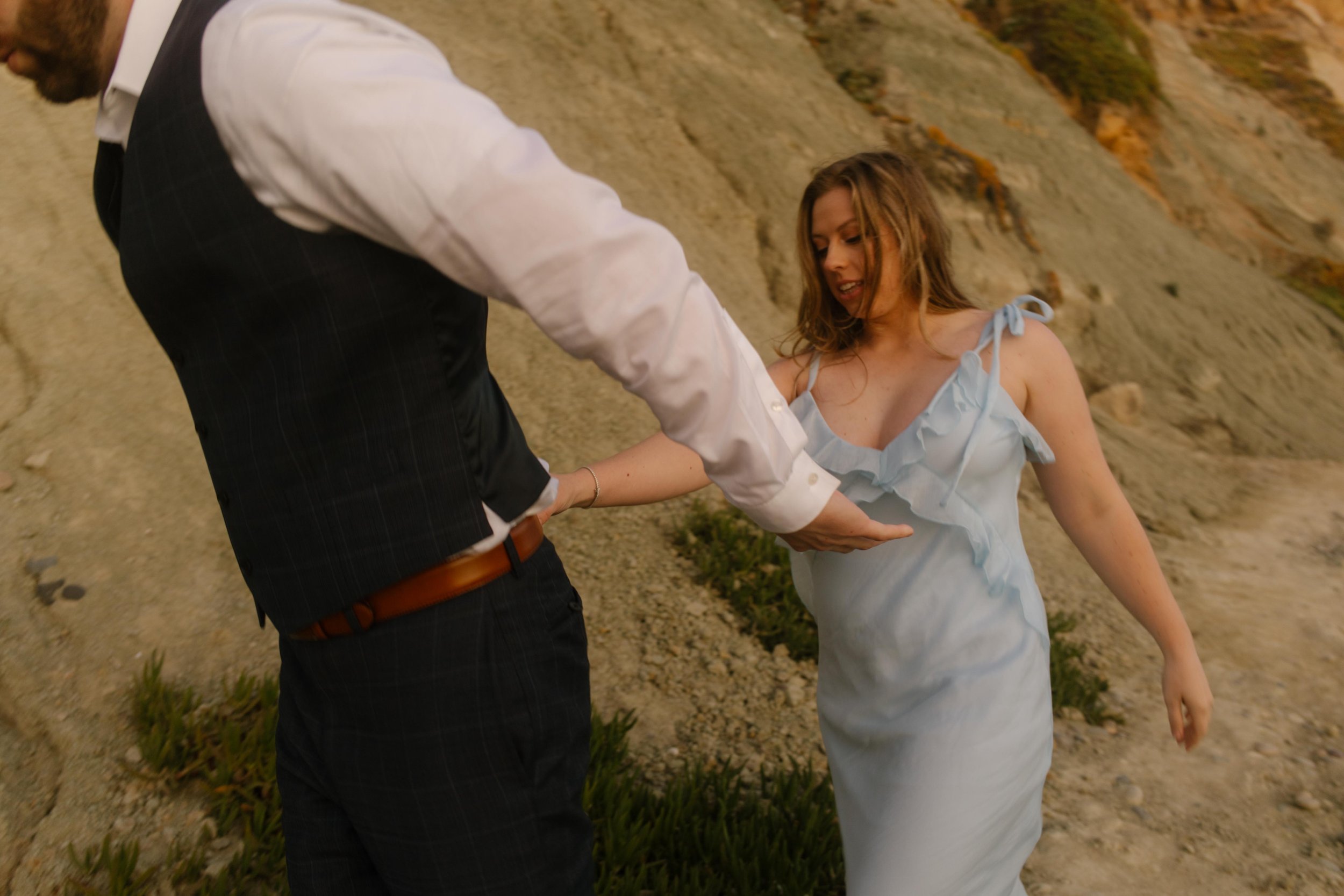He reaches back to help her down the cliffside trail, her soft blue dress flowing with each barefoot step — a candid moment from their San Diego engagement photos.