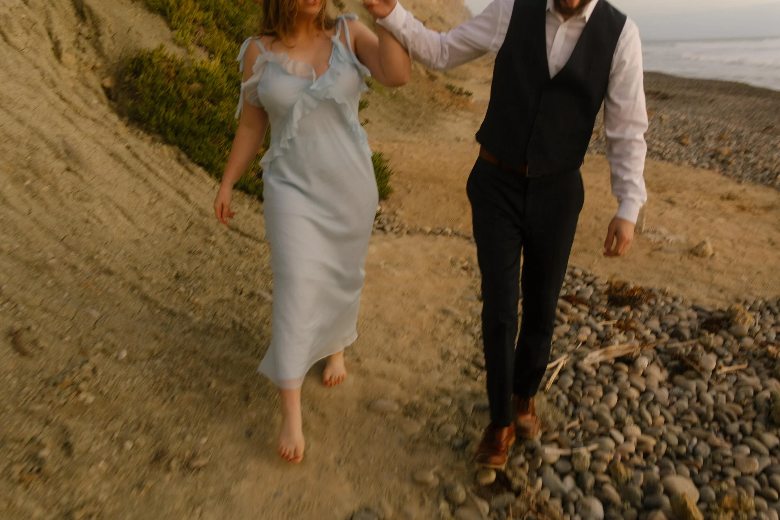 The couple walks hand-in-hand along the shoreline path, her bare feet and breezy dress catching that golden San Diego light.