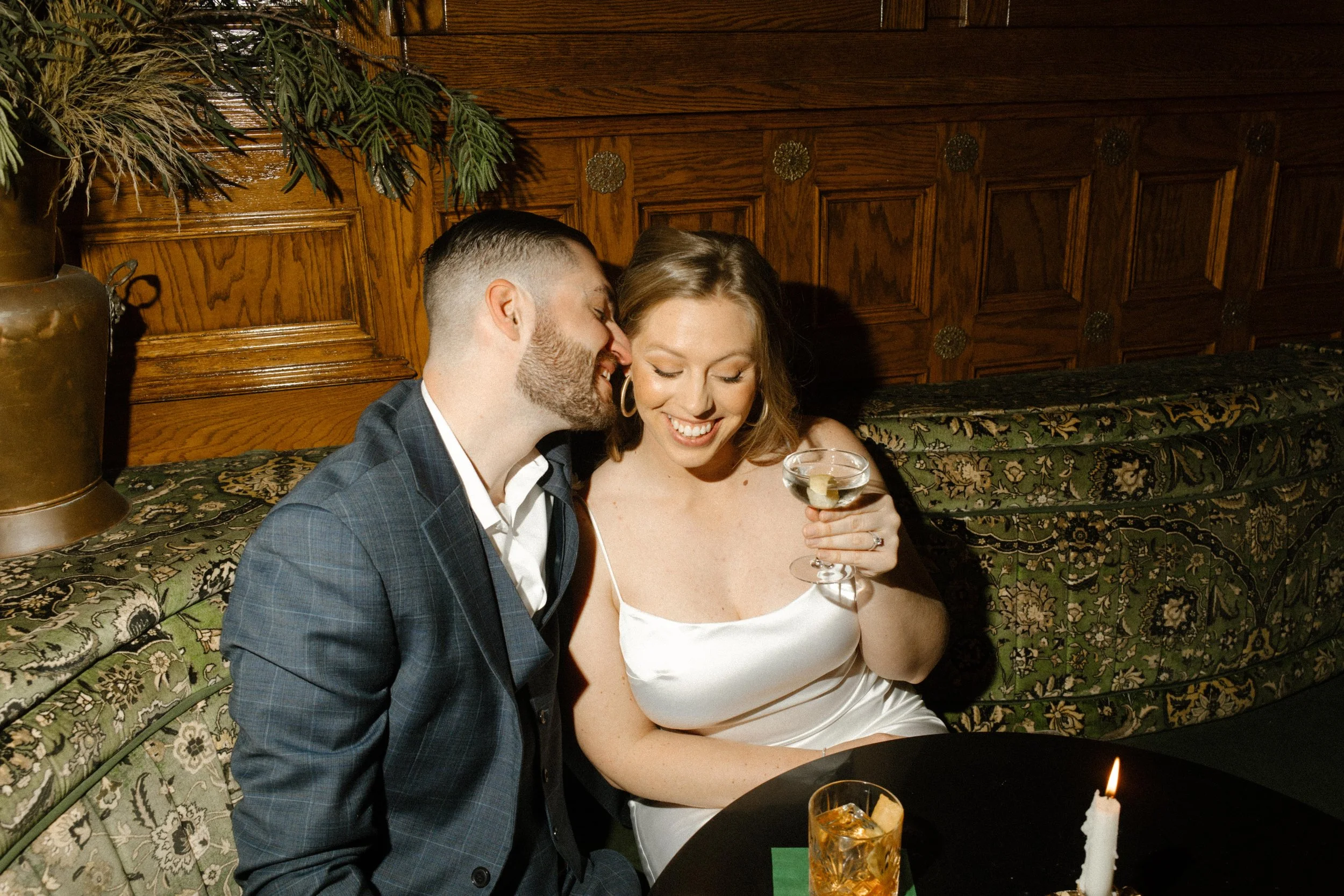 He whispers in her ear while she giggles into her martini glass — cozy vibes and golden-hour glam during their San Diego engagement session.