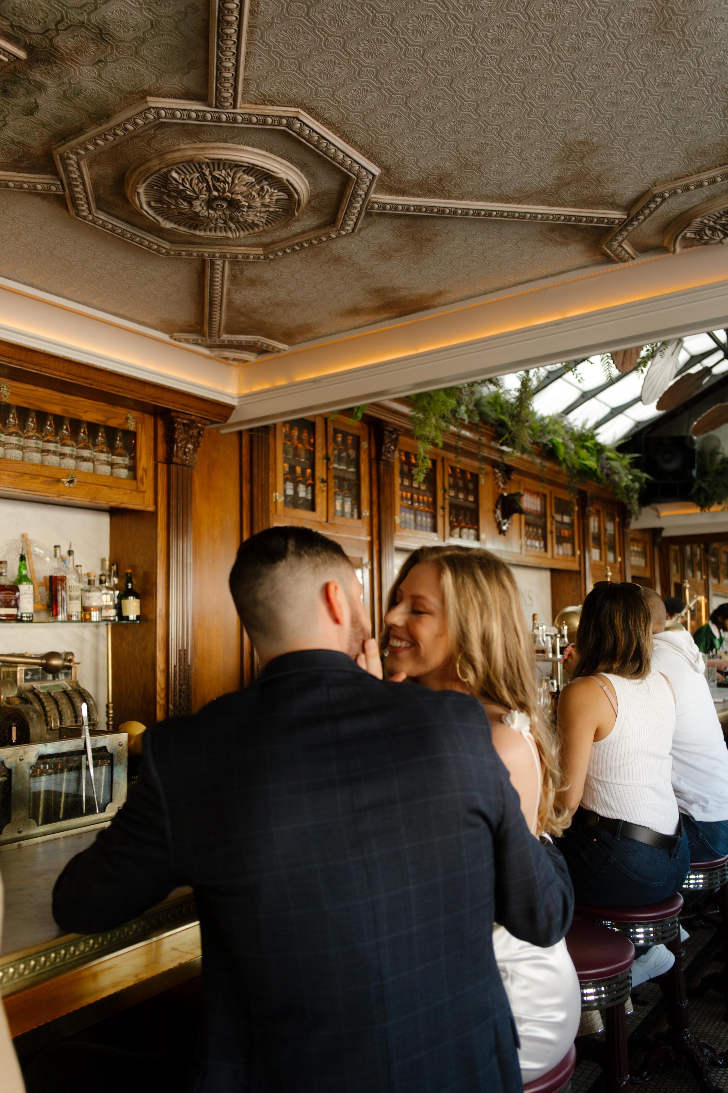 They lean in with quiet laughter at the bar, surrounded by ornate ceilings and soft morning light.