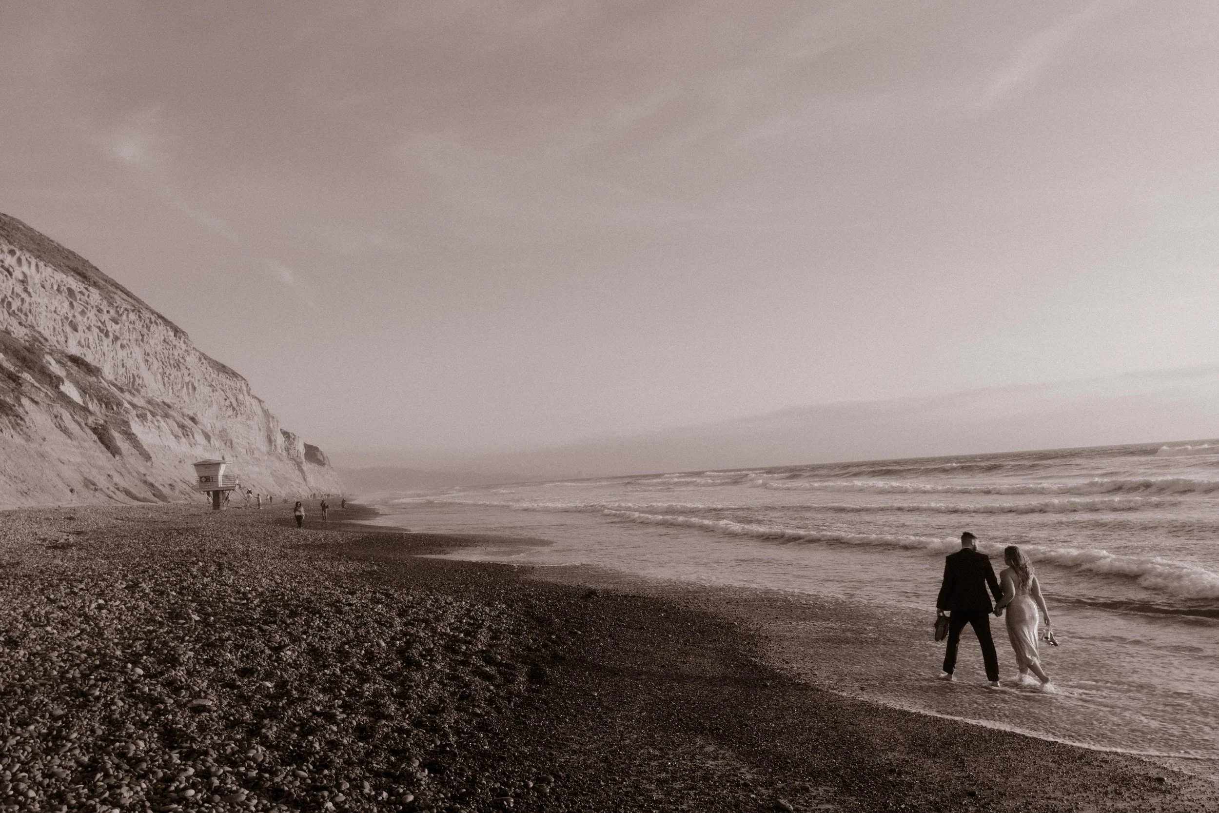Holding hands by the waterline, the couple strolls the beach beneath towering cliffs — timeless and full of quiet connection.