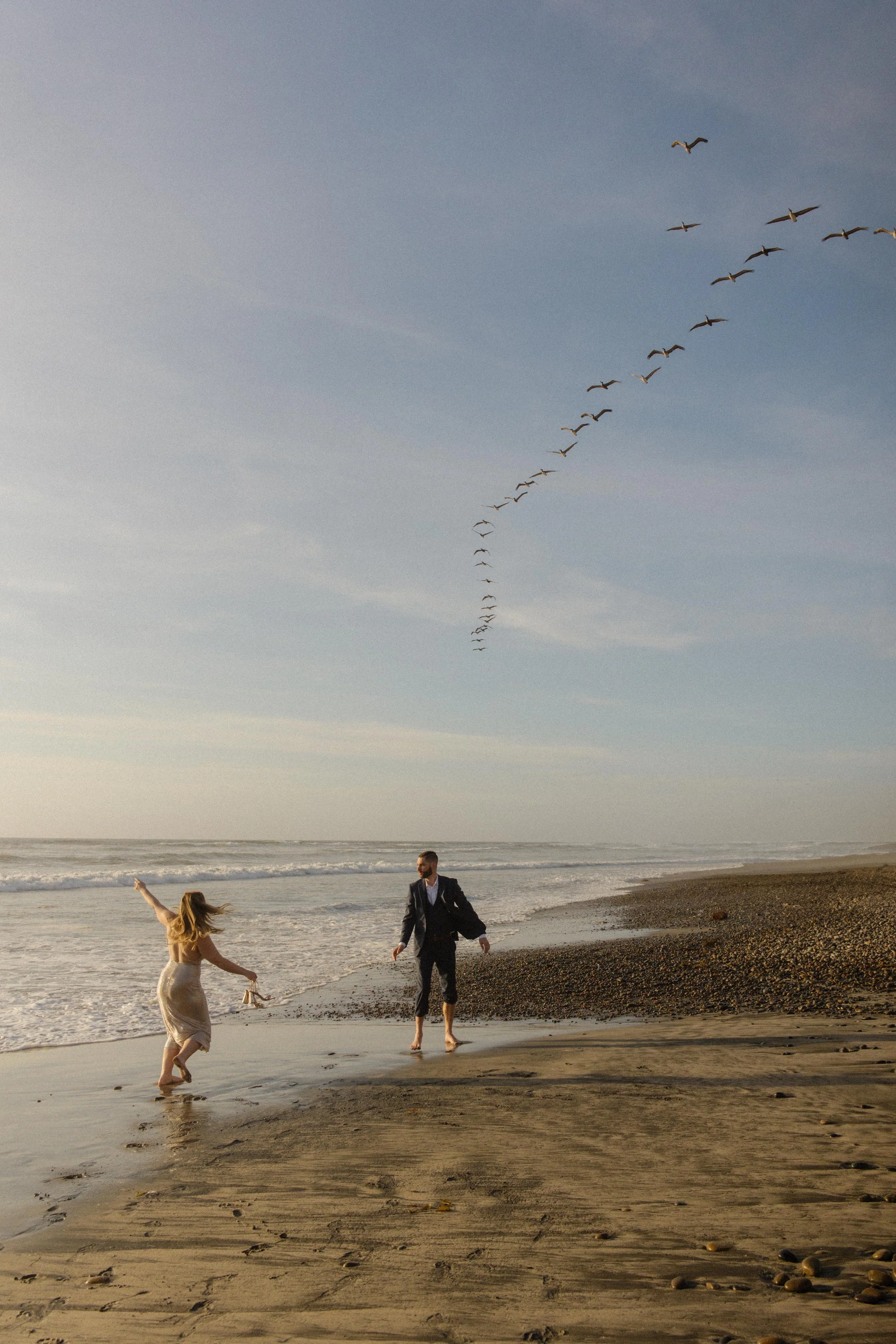 The couple runs barefoot through the surf as a flock of birds flies overhead — wild, joyful, and full of movement.