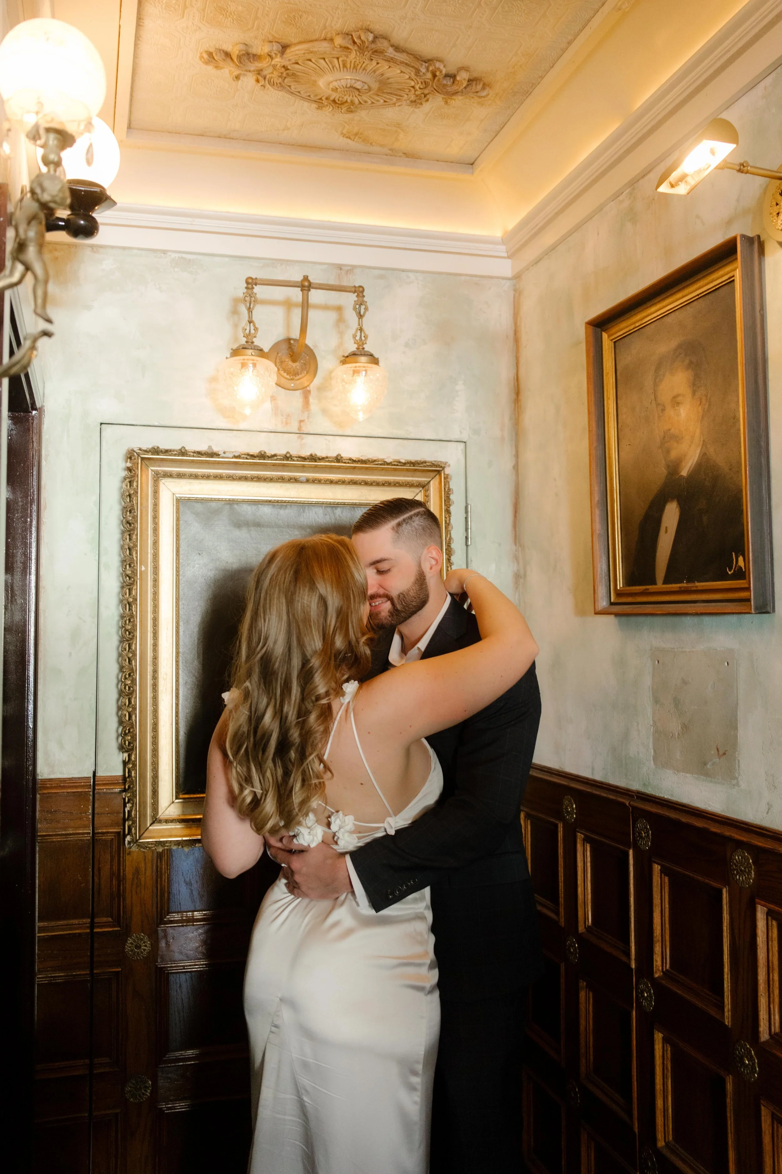 Framed by antique portraits and warm lighting, they lean into each other for a quiet, romantic moment.