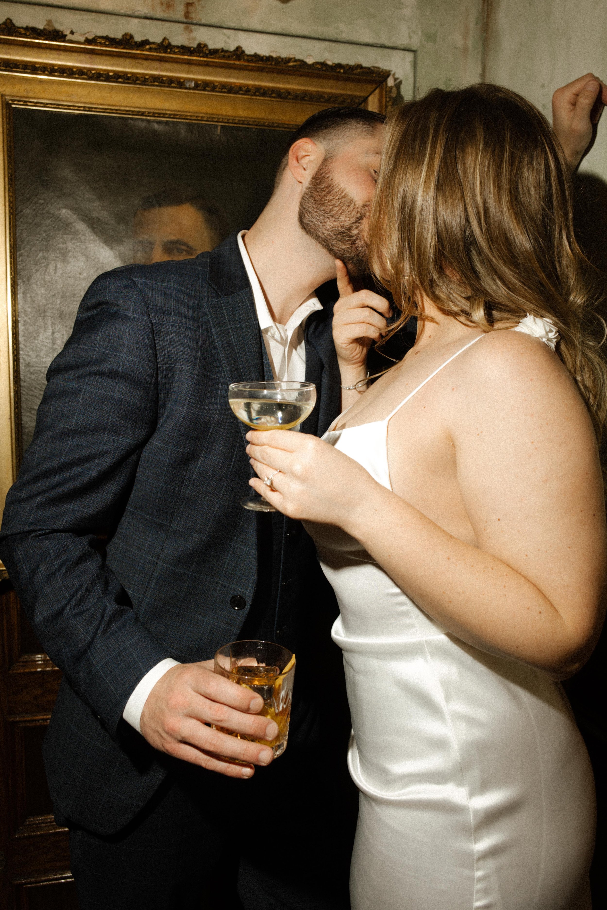 They kiss between sips, both holding classic cocktails — a sultry, stylish bar moment full of heat and chemistry.