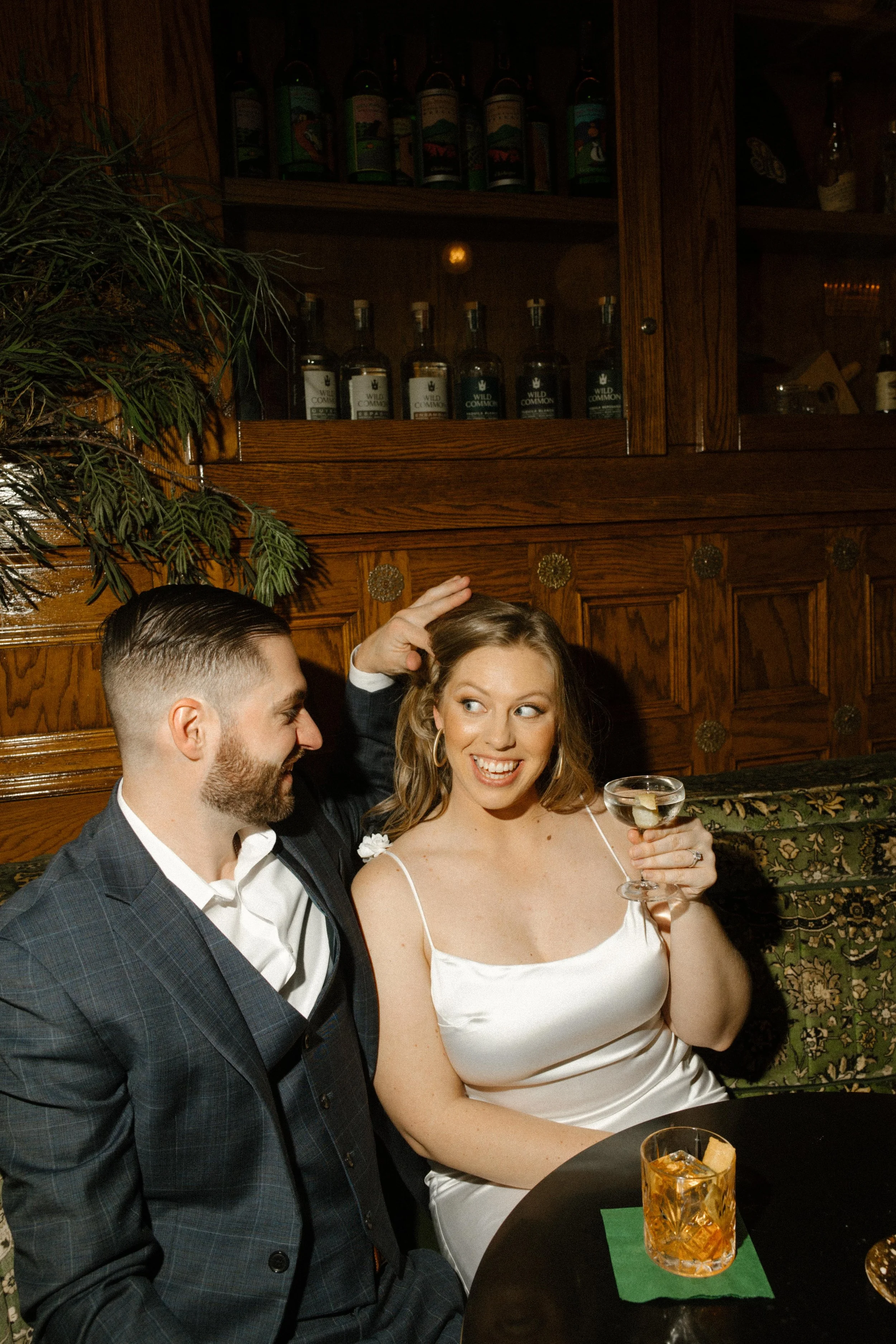 She laughs with a raised brow and a martini in hand while he playfully reaches for her hair — bold and playful energy from their San Diego engagement photos.