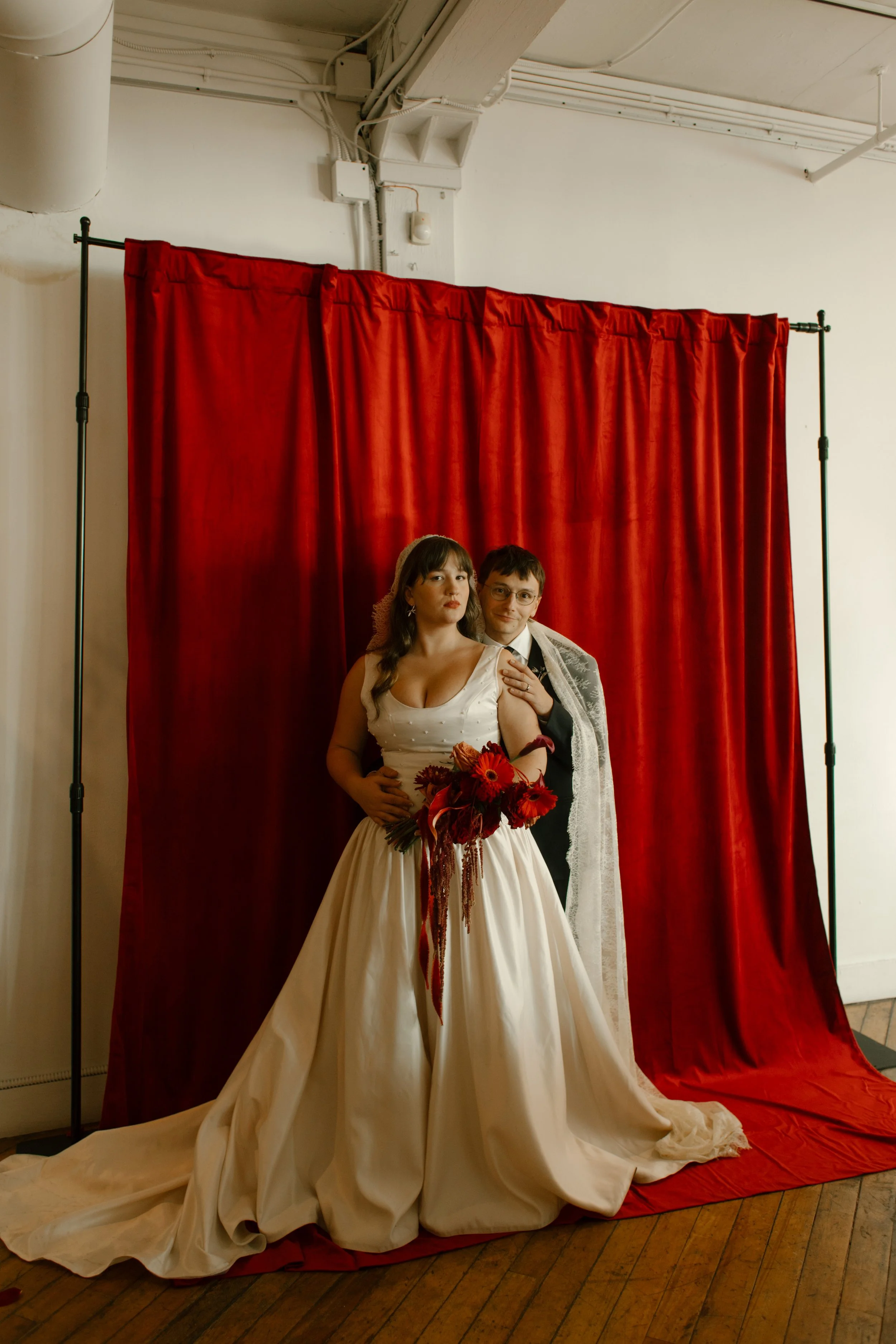 Bride and groom embrace in front of vintage red curtain during indoor bridal portraits