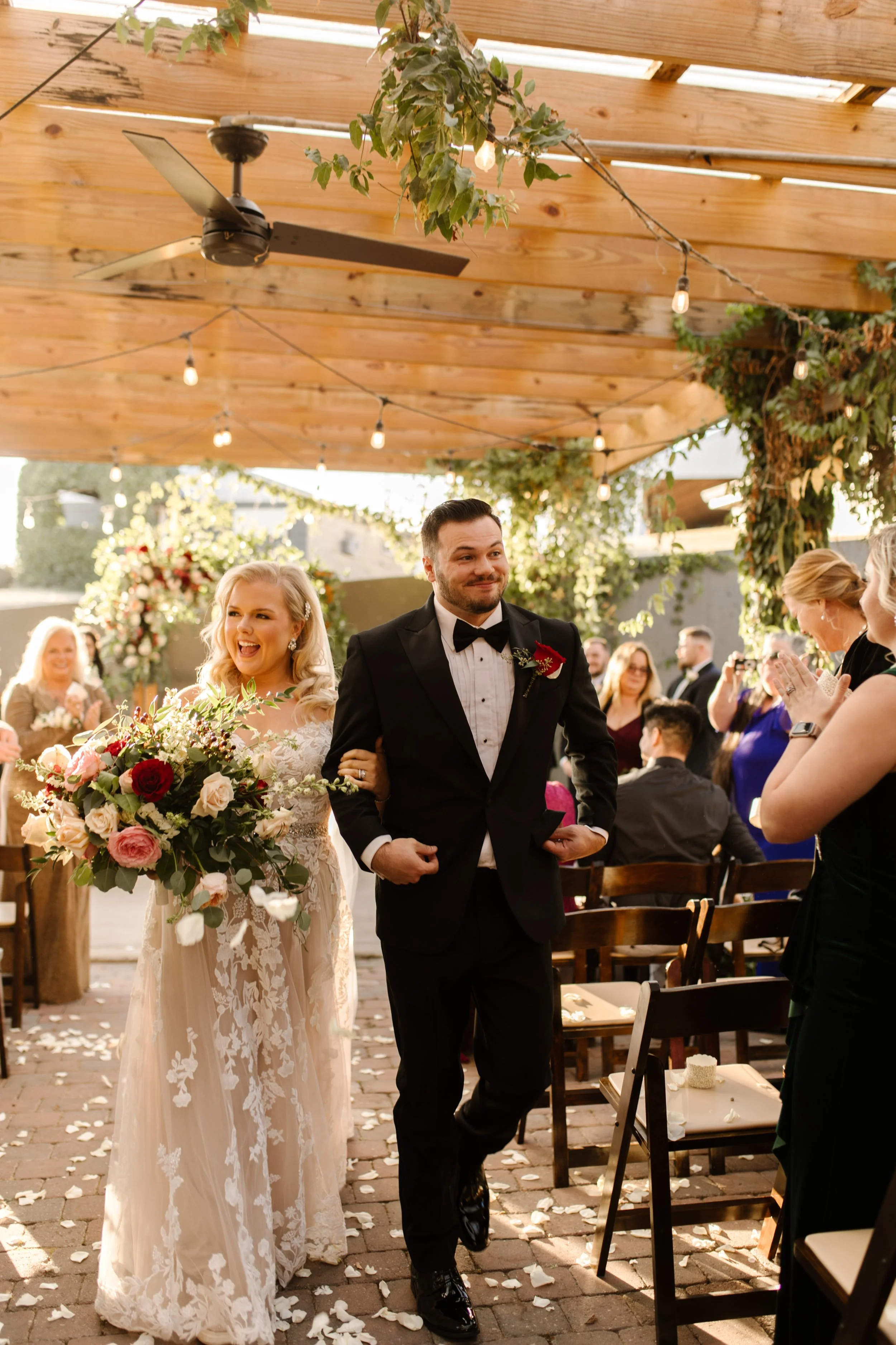 Bride and groom walking down the aisle smiling during their italy inspired wedding ceremony under a wooden pergola with string lights and lush greenery.