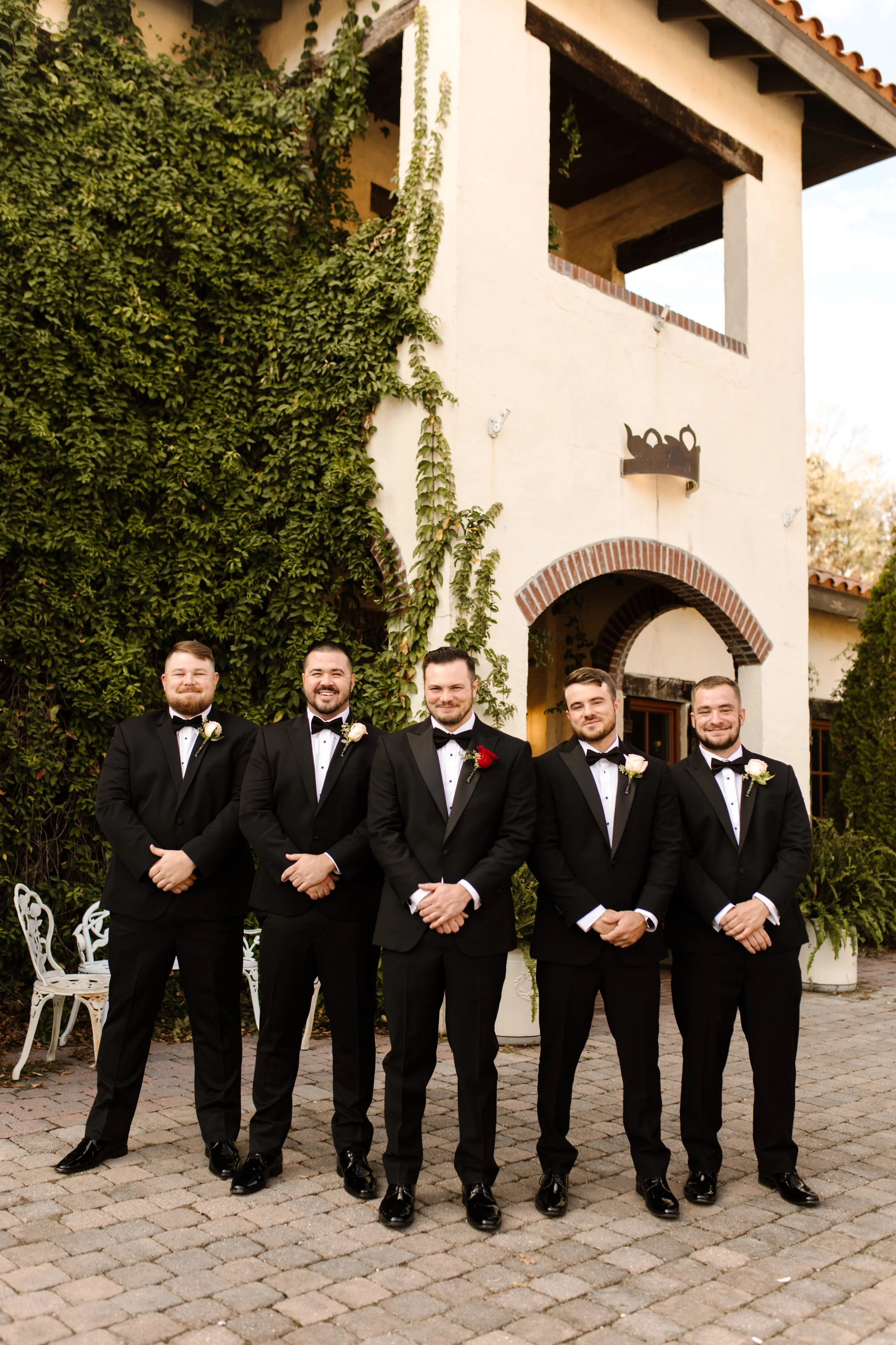 Groom and groomsmen in black tuxedos posing in front of an ivy-covered villa during an italy inspired wedding celebration.
