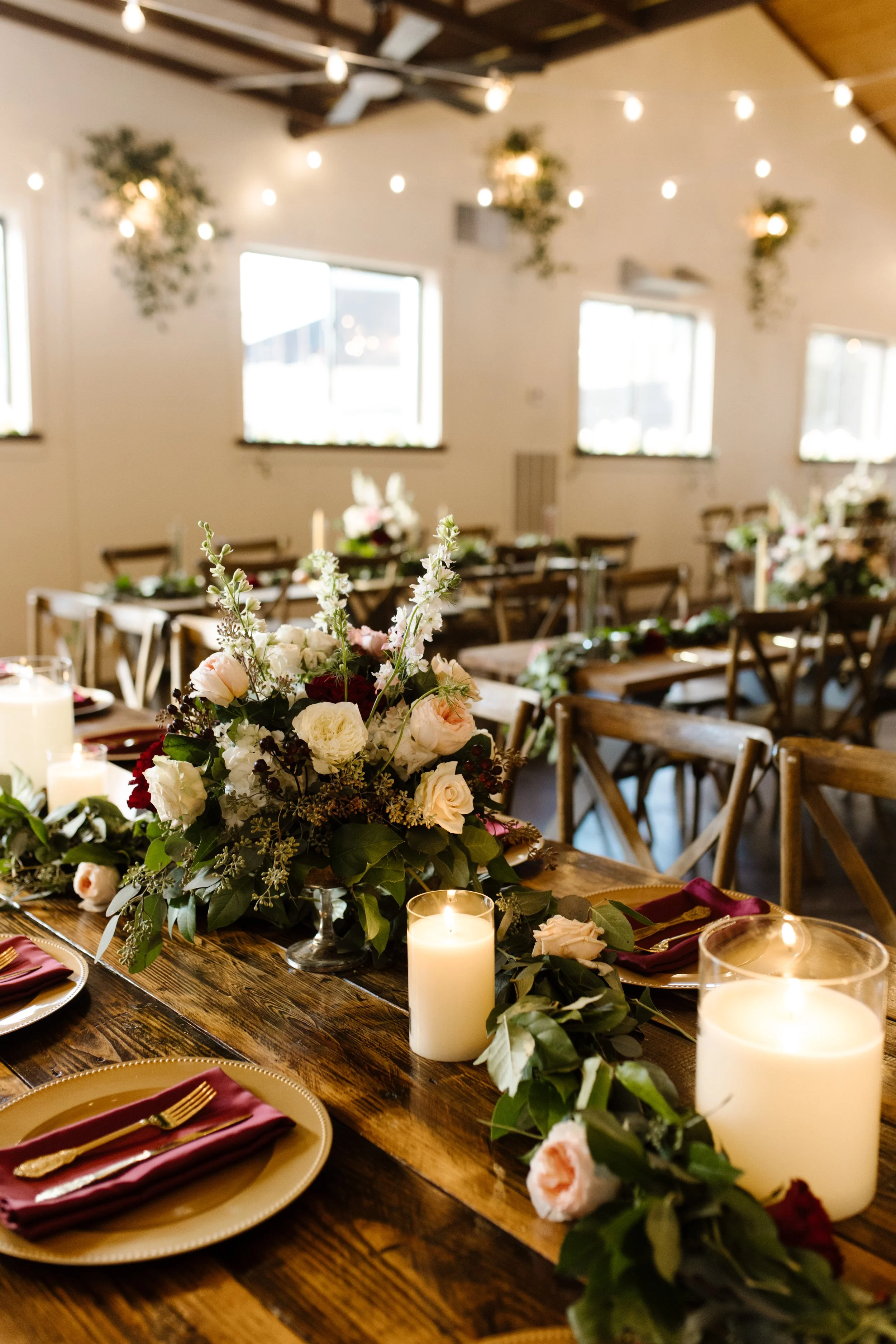Romantic reception table setup at an italy inspired wedding featuring lush floral centerpieces, gold flatware, burgundy napkins, wooden farm tables, and glowing candlelight.
