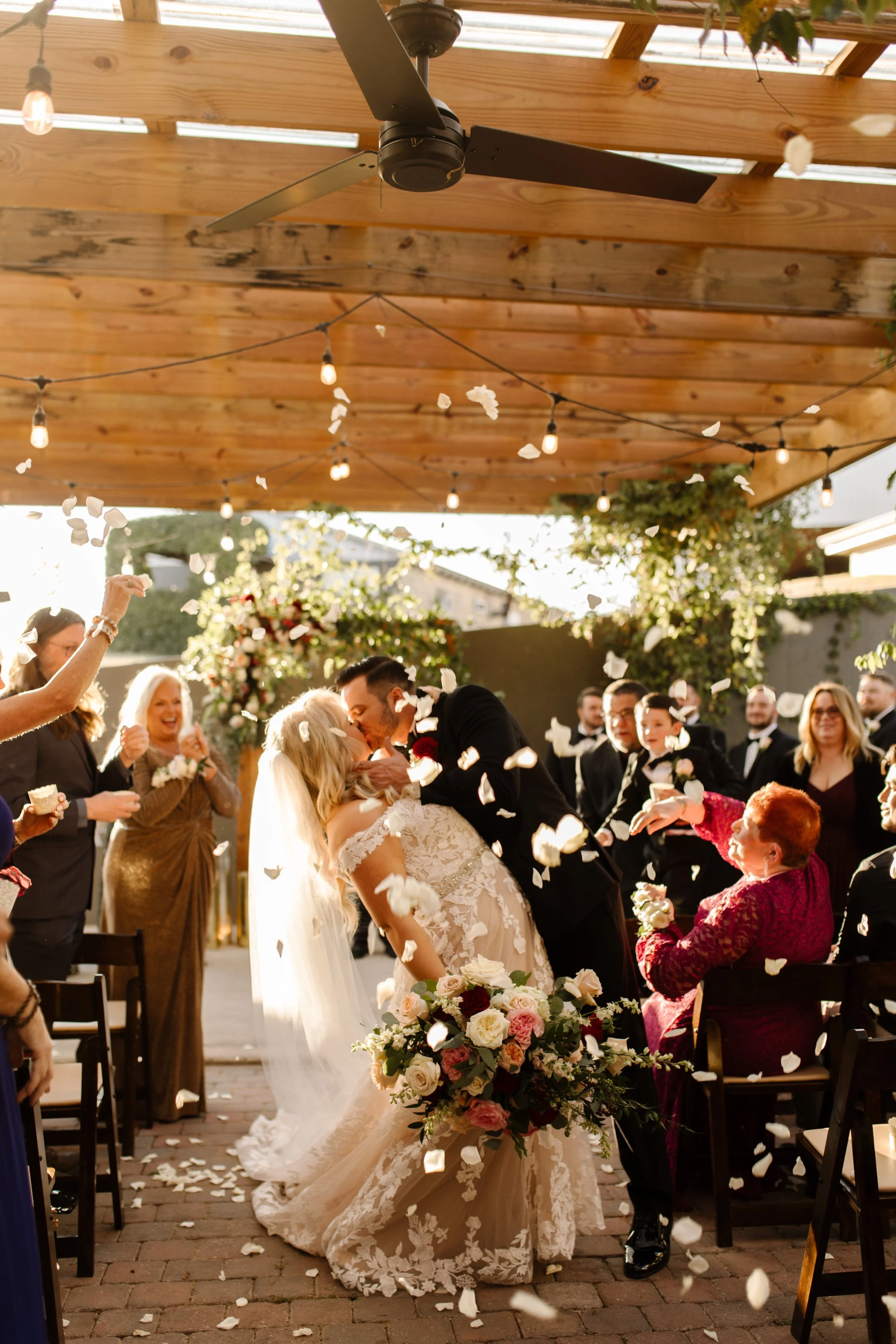Bride and groom sharing their first kiss under a wooden pergola as guests toss petals during an italy inspired wedding ceremony.