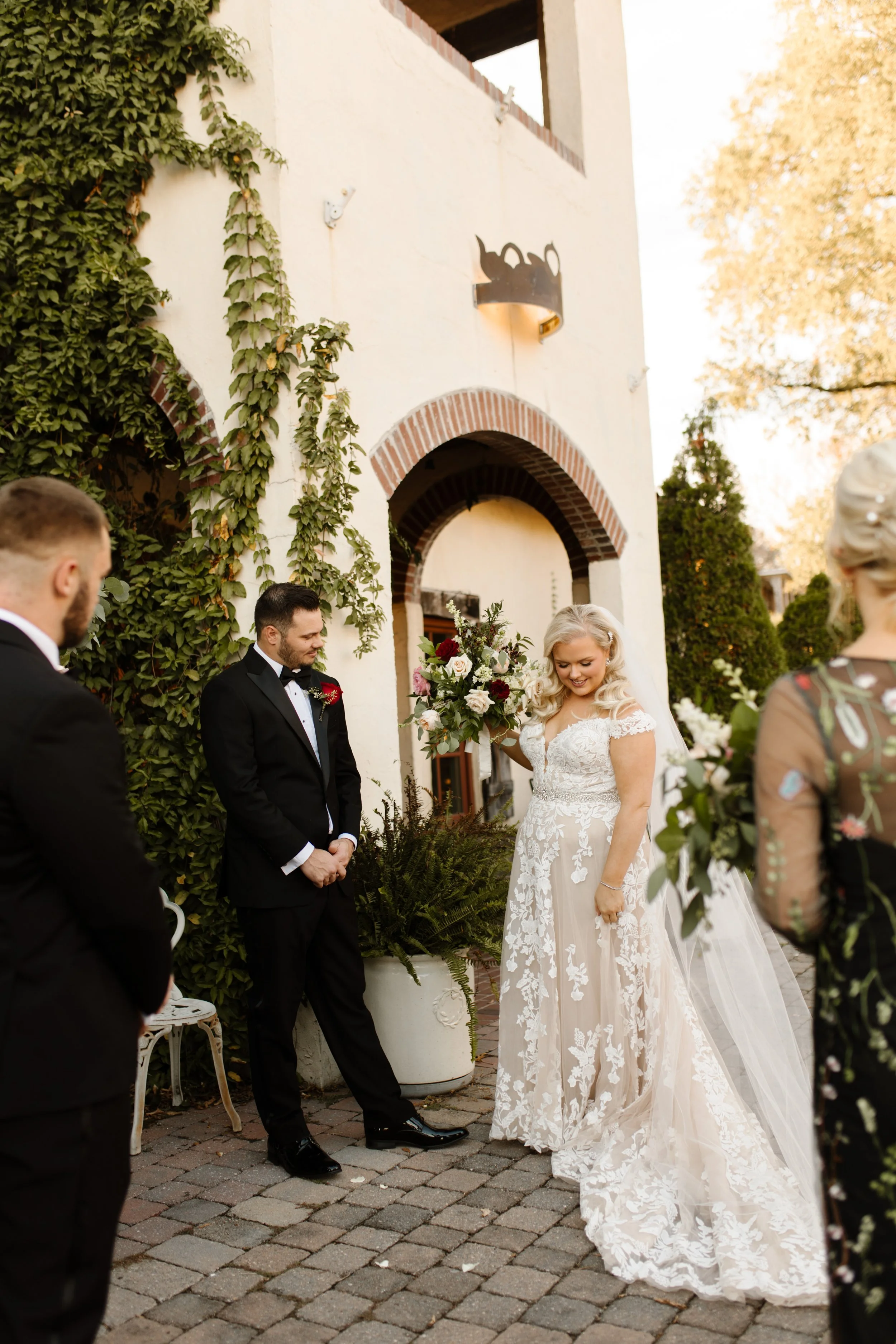 Bride and groom standing outside an ivy-covered villa at their italy inspired wedding, bouquet overflowing with blush and deep red florals.