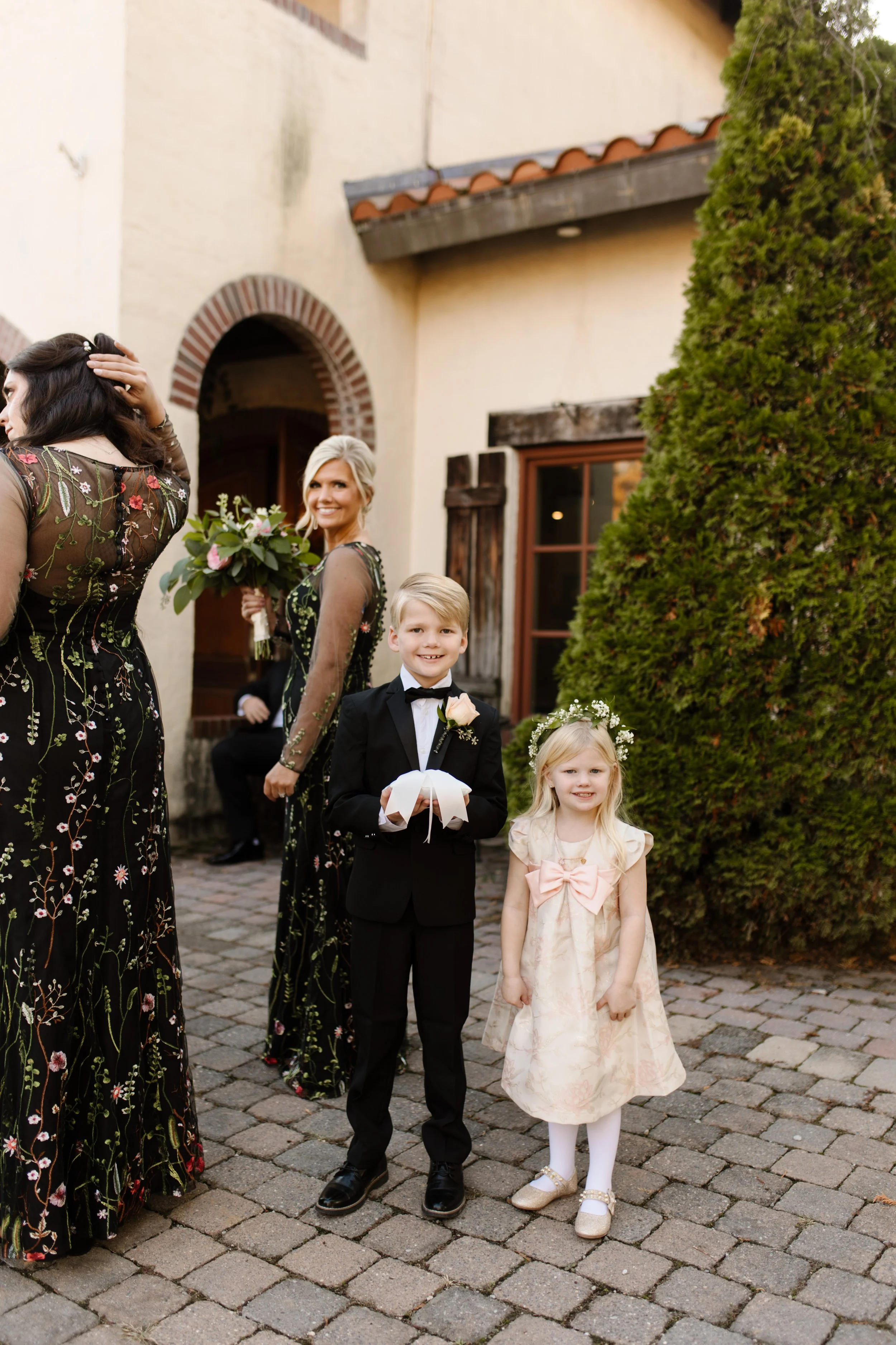 Ring bearer and flower girl posing in front of the villa during an italy inspired wedding with elegant black floral bridesmaid dresses nearby.