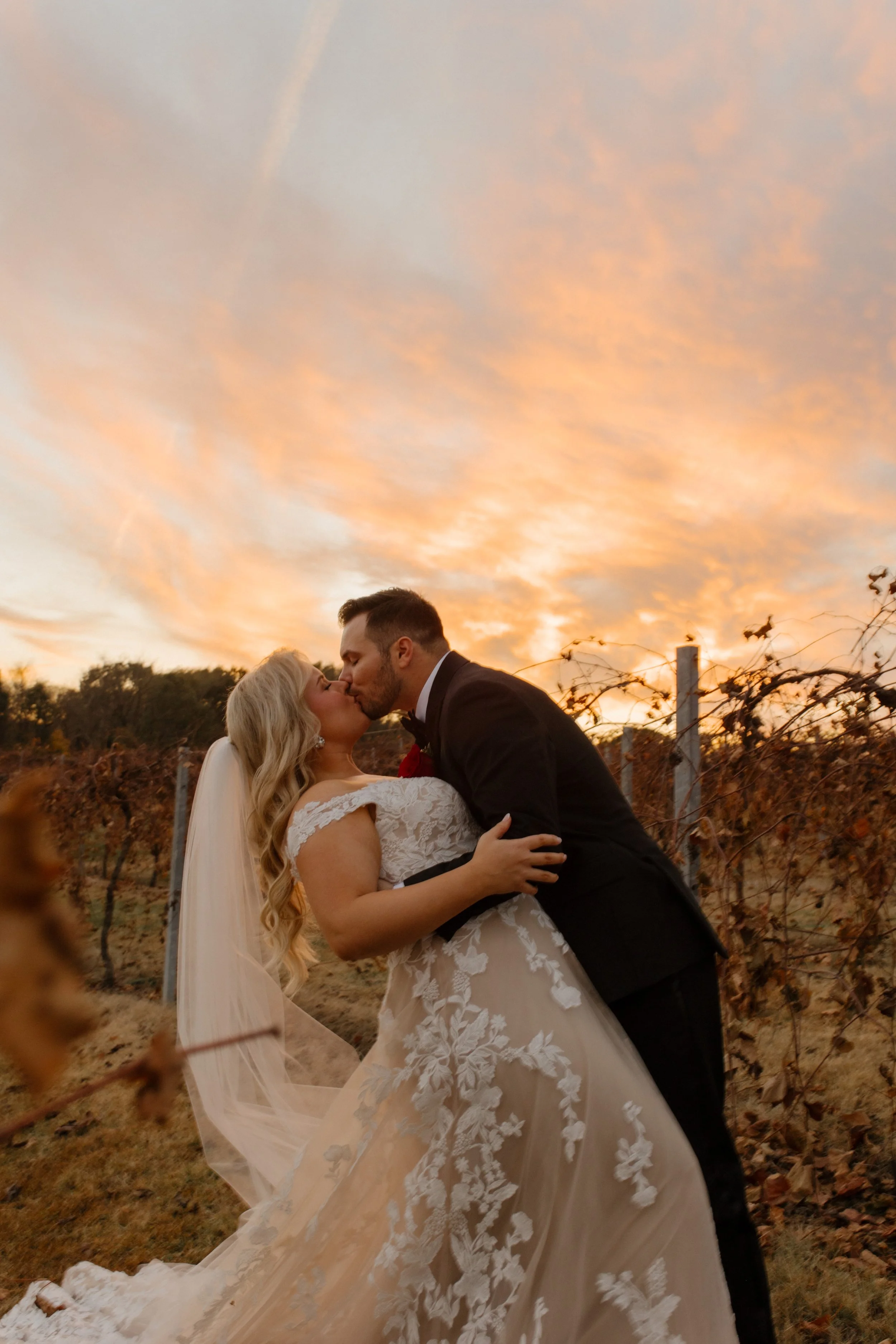 Bride and groom kissing in the vineyard at golden hour during their italy inspired wedding with dramatic sunset clouds overhead.