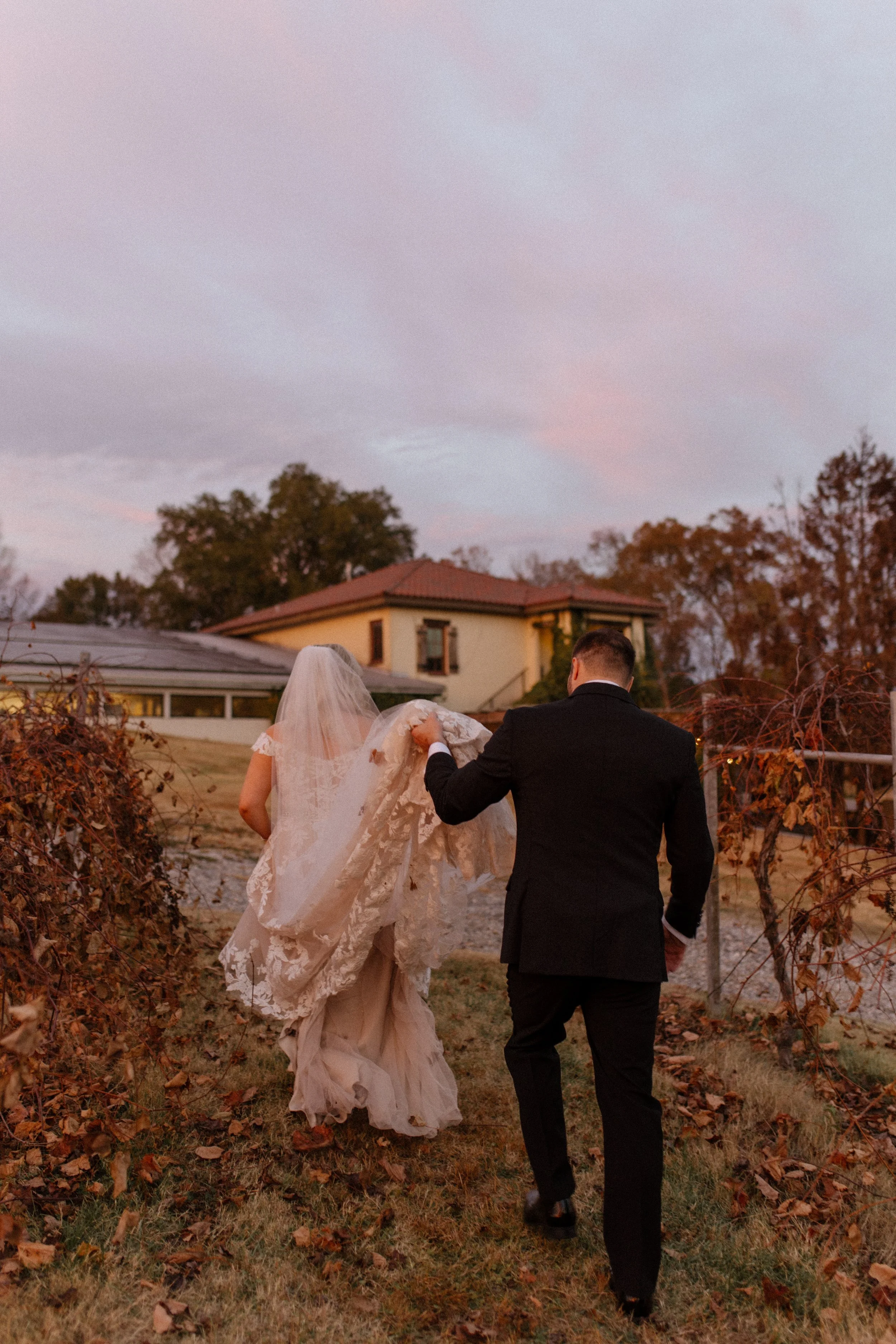Bride and groom walking through vineyard rows at sunset during their italy inspired wedding, surrounded by autumn leaves and soft pink skies.