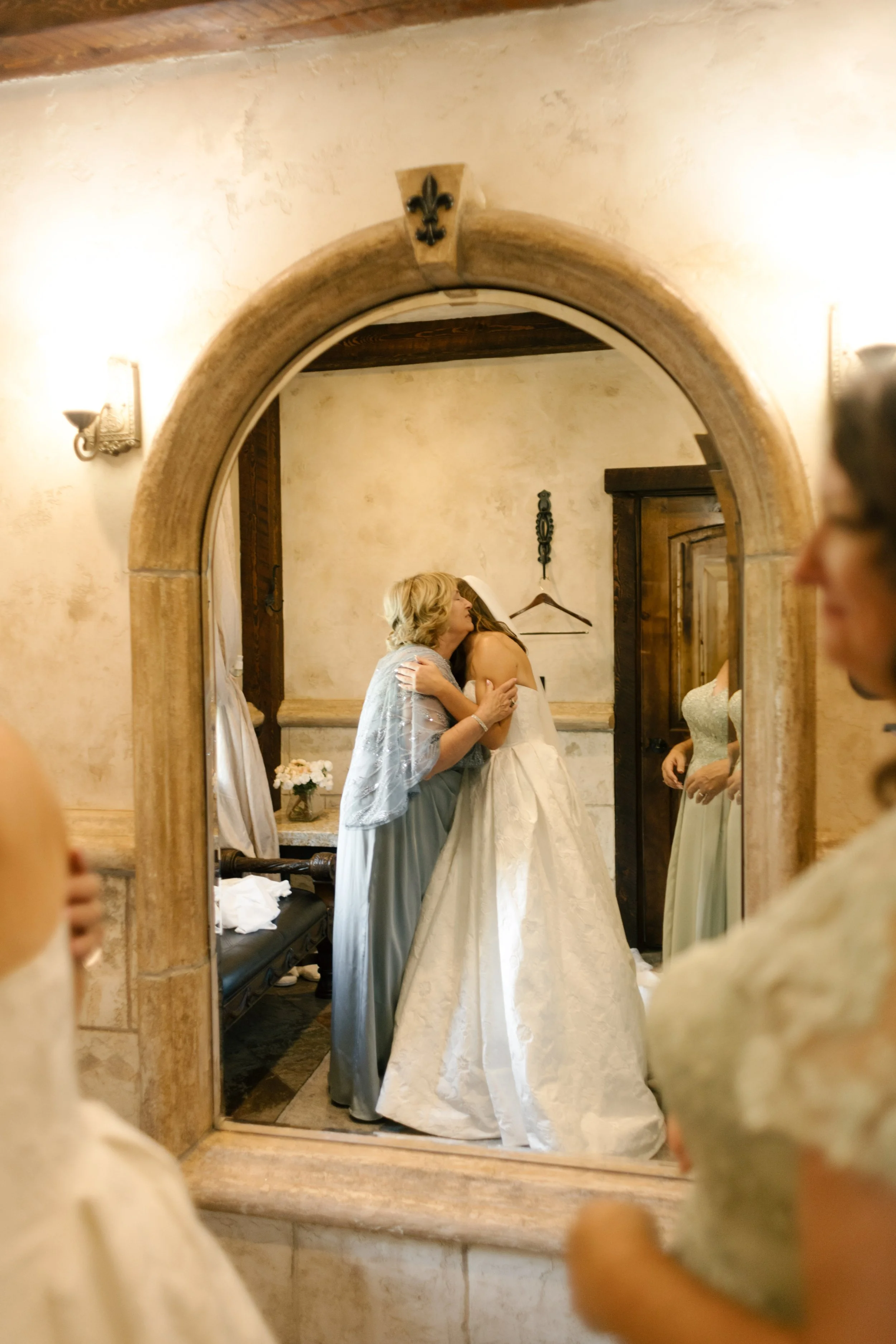 Mother of bride helps bride into her dress and hugging her from behind.