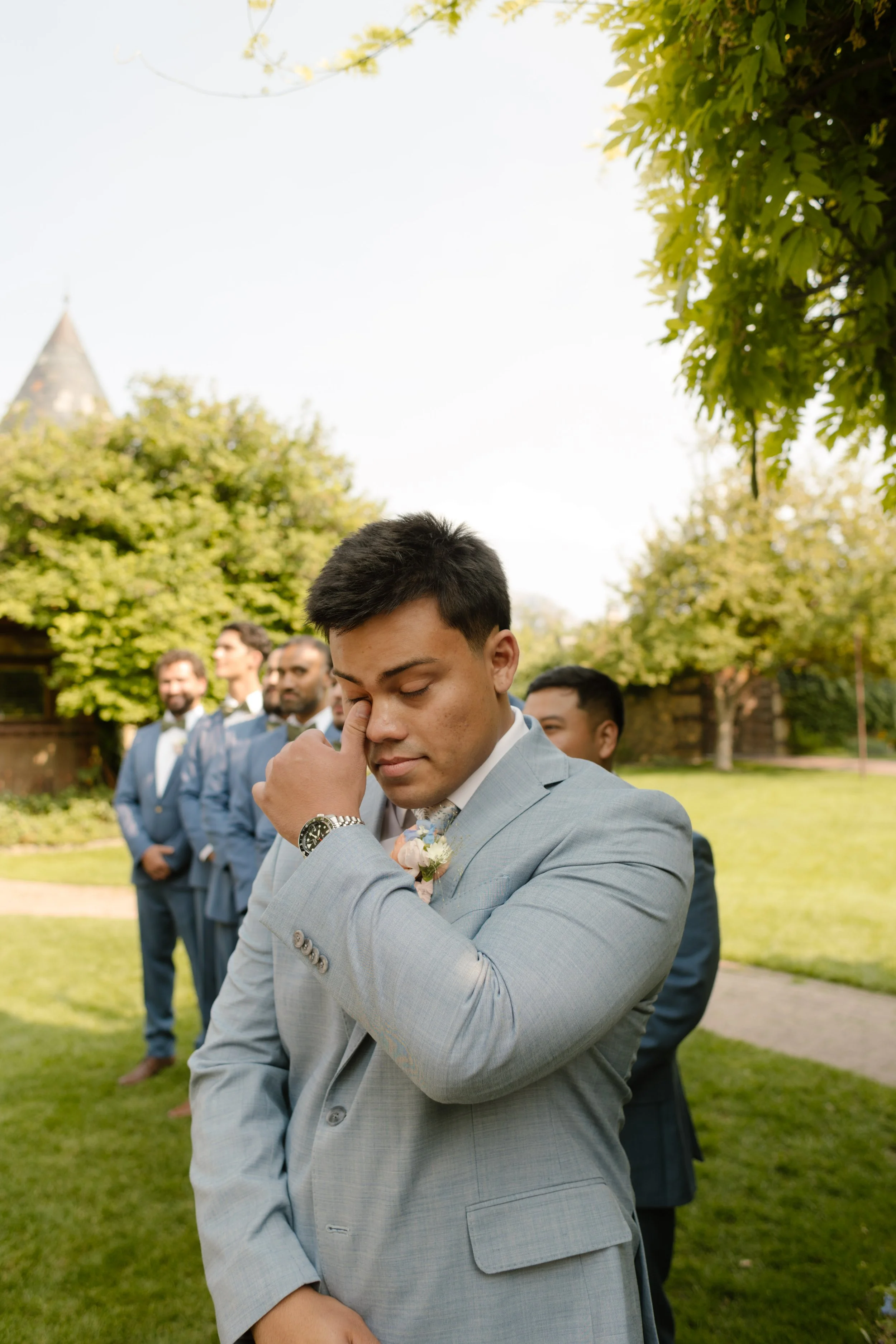 Groom wiping away tears while standing with his groomsmen on a sunny lawn at an outdoor utah wedding venue.